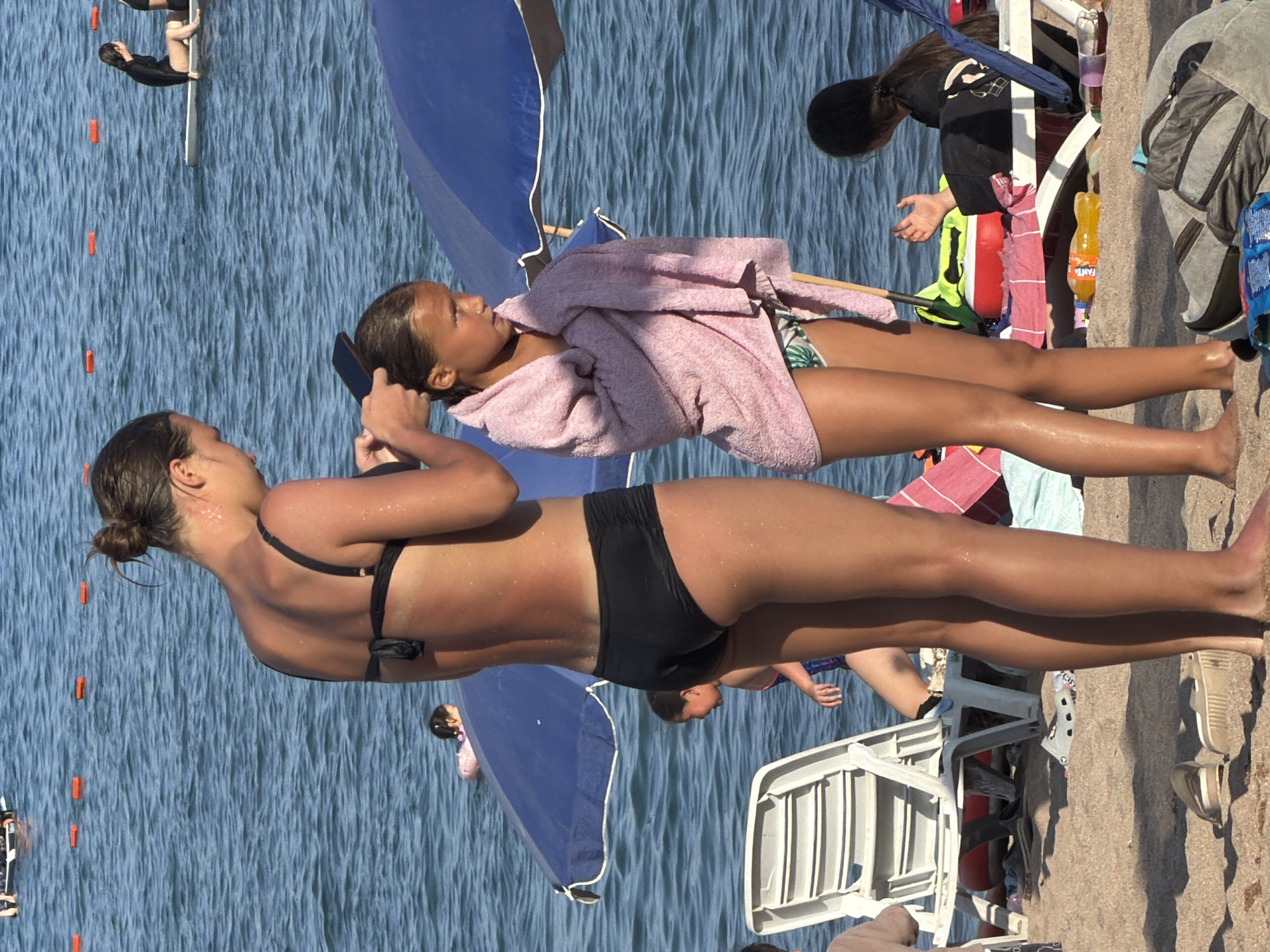 The media file shows a group of people at a beach. The scene is viewed from a high angle, looking down at the subjects. In the foreground, two girls are lying on the sand, one wearing a pink towel and the other in a black bikini. Their legs are in focus and their bodies are partially visible. Behind them, a blue umbrella is set up on the sand. Further in the background, a few other people are visible, including a person wearing a black shirt and a person in a black swimsuit who appears to be climbing out of the water. The water is blue and appears to be calm. The sand is light brown. There are some beach chairs and bags scattered around. The lighting suggests it is daytime, likely sunny, as indicated by the bright lighting and shadows. No specific emotions are overtly displayed by the subjects, but the overall atmosphere appears to be relaxed, characteristic of a beach day. There is some visible text on a bottle of Fanta in the background.