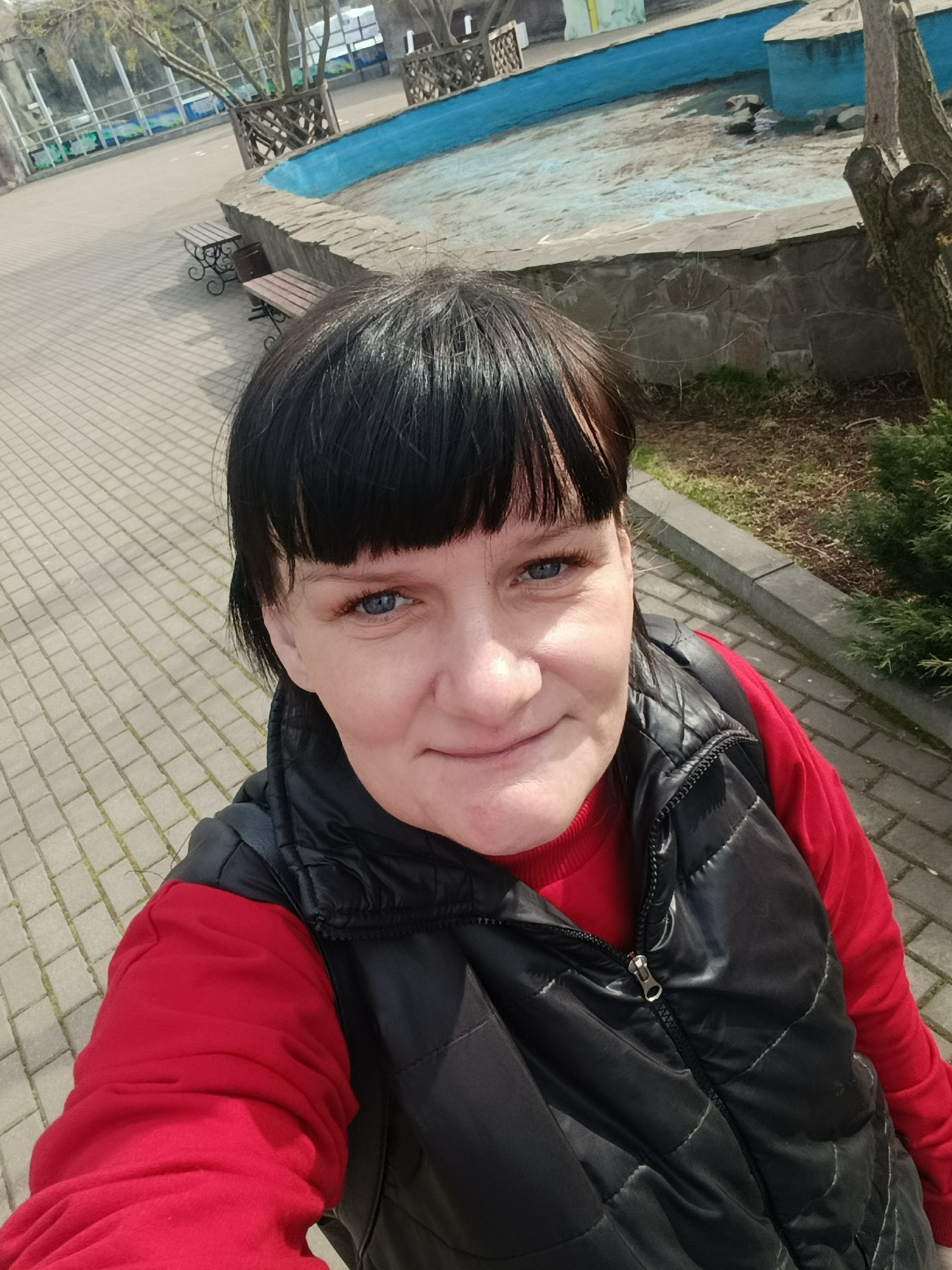 A woman with dark hair and bangs, and distinct blue eyes, is captured in a close-up selfie. She wears a bright red long-sleeved top under a black quilted vest with a zippered front, and has a slight, pleasant smile.
The setting is an outdoor public space, likely a park or garden in Izhevsk, Russia. She is standing on a paved area made of light-colored interlocking bricks. In the background, to her left, are two small wooden benches with black ornate metal bases, positioned near a dark cylindrical trash can. A low wall constructed from grey, irregular stones forms the edge of what appears to be a dry, shallow fountain or pool. The interior of this structure is painted a light blue, showing signs of wear and some debris.
Beyond the dry fountain, there are bare tree branches, indicative of an early spring or late autumn season. Green bushes and patches of natural ground are also visible. The bright, even lighting suggests it is daytime with sunny conditions. No visible text is present in the image.