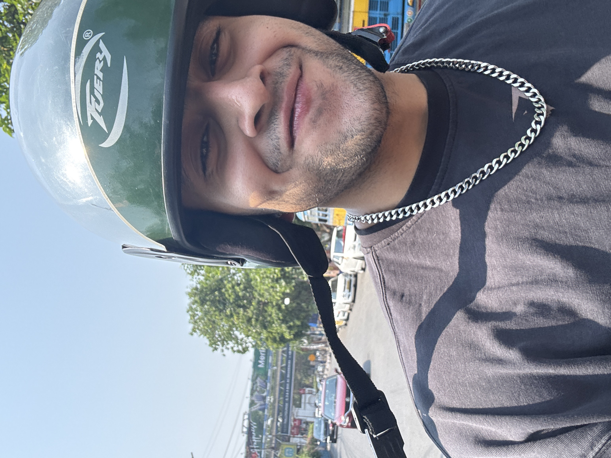 A selfie of a light-skinned man wearing a green helmet with the logo "Tuery" and a silver chain around his neck. He is wearing a dark t-shirt. The background shows a city street, and a sign that reads "Merlin" suggests that the location is Kolkata, India. Several cars are visible. The sky is clear.