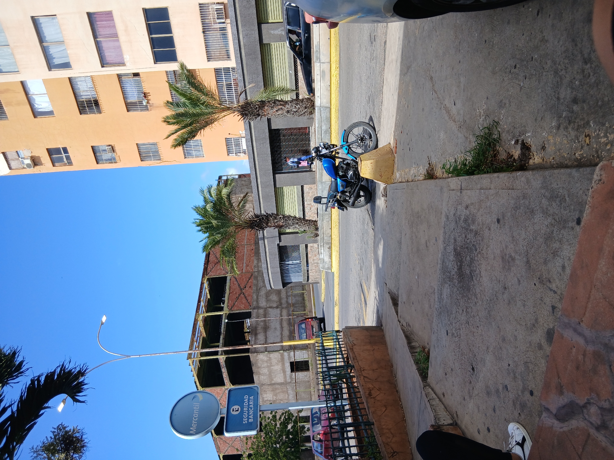 The image shows a street scene in Juan Griego, Venezuela. In the foreground, a concrete sidewalk curves to the right. To the left, a person wearing black pants and white sneakers is partially visible, seated on a red brick step. A blue motorcycle is parked on the street, facing left, with its kickstand down. Behind the motorcycle, a person in a blue shirt and dark pants walks away from the viewer. Further down the street, a black car is parked. In the background, there are two palm trees and a modern building with multiple stories and many windows. To the left of the building, there is a sign with the word "Mercantil" and a smaller sign that says "SEGURIDAD BANCARIA" with an "E" inside a circle. Another building under construction, with exposed red brick and metal framing, is visible. A street light with two lamps stands to the left of the frame. The sky is clear and blue.
