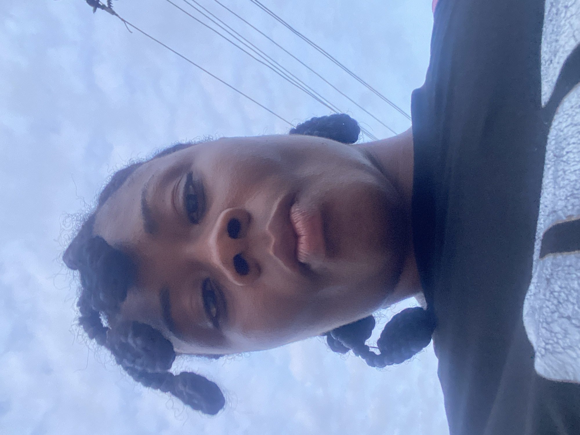 A person is looking down at the camera from a low angle. The person has dark hair styled in braids, and is wearing a dark shirt with some white pattern on the side. The sky is visible in the background, with several wires overhead. The location cannot be reliably determined as Lagos, Nigeria.