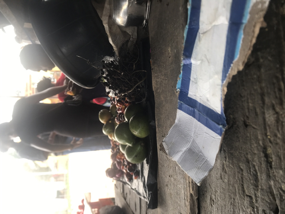 The image shows a street vendor selling fruit in Umudike, Nigeria, located at latitude 5.481 and longitude 7.546. The vendor's stand is located on a wooden plank and consists of a variety of fruits including green mangoes and palm fruits. The vendor is obscured from view, but several customers are visible, some with their hands outstretched, presumably towards the fruit. The foreground consists of a close-up of the fruit stand, with the remainder of the scene out of focus. To the right of the fruit stand is a piece of cardboard with blue and white stripes. There is an empty space to the left of the fruit stand that reveals the street beyond. The scene appears to be lit by sunlight from above.
