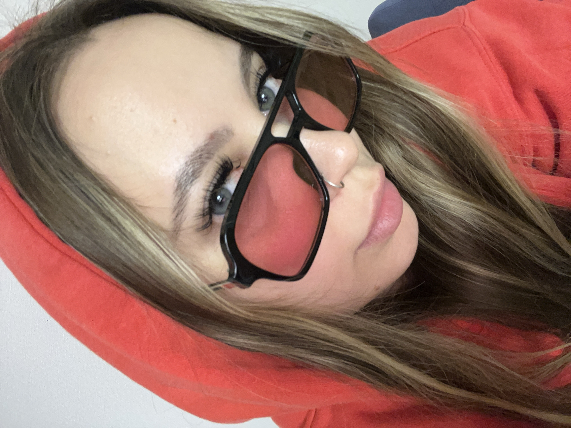 The image is a close-up, angled portrait of a young woman looking directly at the viewer. Her head is tilted, and she is wearing a bright coral or reddish-orange hooded sweatshirt, with the hood up to frame her head. Her long, wavy brown hair with lighter highlights flows around her shoulders and over the hoodie.
She wears black-framed sunglasses with reddish-tinted lenses, which are pushed down on her nose, allowing her light blue eyes to be clearly visible above them. Her eyes feature long, dark eyelashes and well-defined eyebrows. A small silver hoop nose ring is visible on her right nostril. Her lips are slightly parted, showing a natural pinkish hue, and her expression is direct and composed.
The background is minimal, featuring a plain light-colored surface on the left and bottom, and a dark blue or grey object behind her head on the right. The lighting is soft and even, illuminating her face without harsh shadows. No text, specific time of day, weather conditions, or explicit location identifiers are visible within the image.