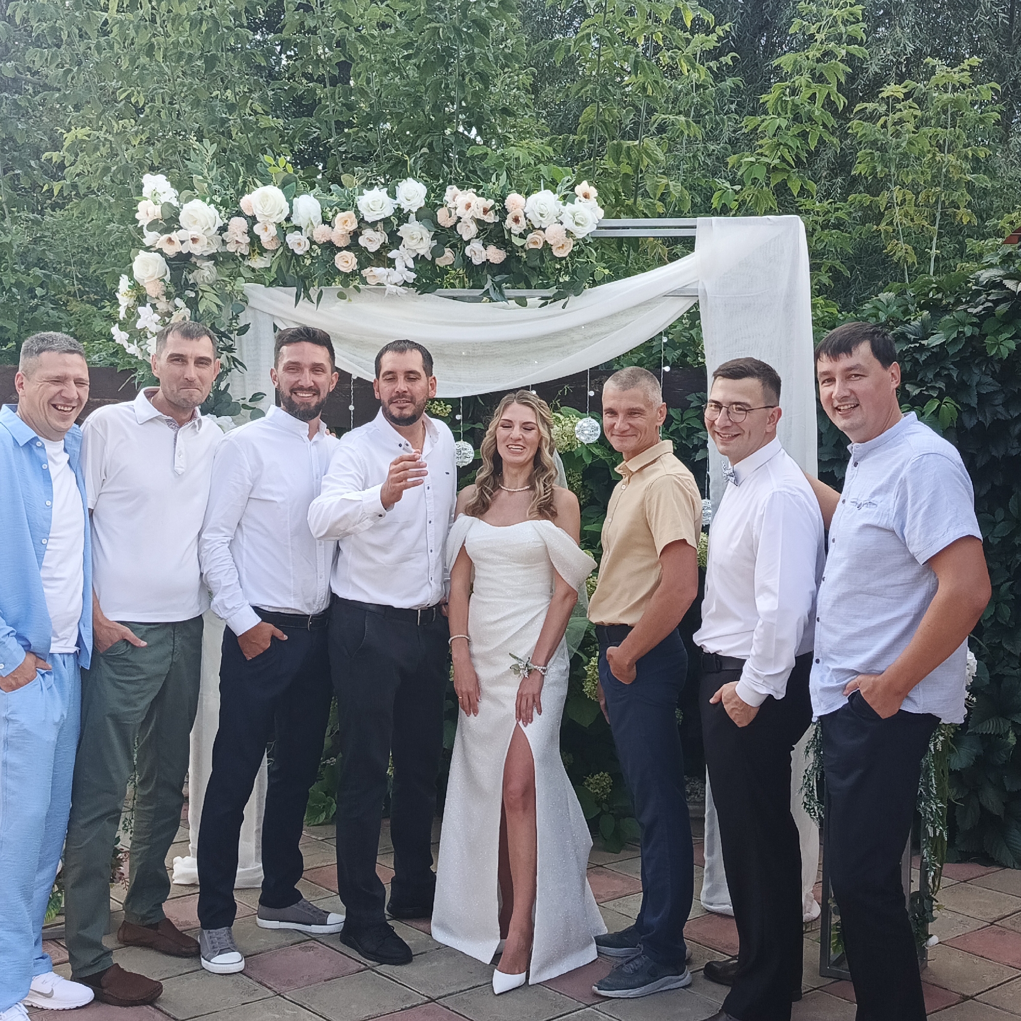 In the city of Chaikovsky, Russia, at an outdoor wedding ceremony, a group of eight people are posing for a photograph in front of a floral archway. Seven men, dressed in various casual and semi-formal attire, stand on either side of a bride in a white wedding gown with a thigh-high slit. The bride stands slightly off-center, smiling broadly, with her hair styled in loose waves and adorned with a veil. The men are all looking towards the camera, with varying expressions of joy and amusement. One man on the far left is wearing a light blue unbuttoned shirt and pants. The others are in white shirts and dark pants, or light colored shirts and dark pants. The floral archway, decorated with white and peach roses, is draped with white fabric and illuminated by string lights. The background consists of lush green foliage, suggesting a garden setting. The ground is paved with reddish-brown and grey tiles.
