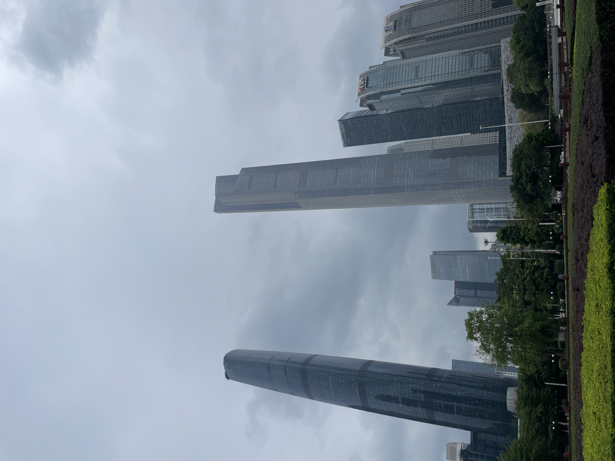 The image depicts a cityscape dominated by tall, modern skyscrapers against an overcast sky. The location is likely in the Tianhe District of China, known for its contemporary architecture. Two prominent structures are the Guangzhou CTF Finance Centre, a cylindrical tower, and the Guangzhou International Finance Center, a rectangular tower. Green foliage and trimmed hedges are visible at the bottom, suggesting a landscaped park or urban garden in the foreground. Lamp posts line the edge of the greenery. The overall environment conveys a sense of urban density and development in a large metropolis. There are no people visible in the image.
