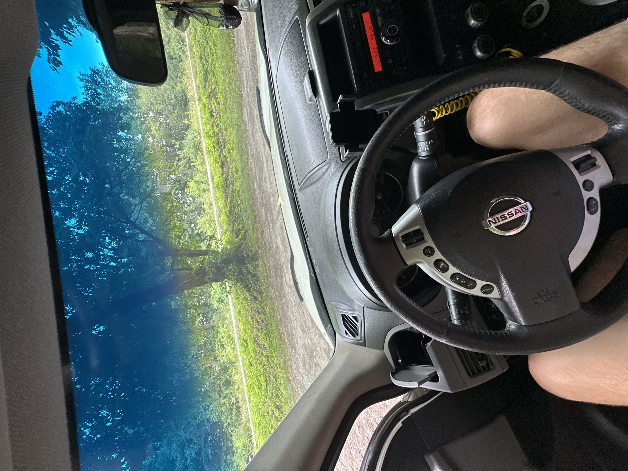 An interior shot of a Nissan car shows a person's leg in the driver's seat, their knee visible to the right of the steering wheel. The steering wheel itself, bearing the Nissan logo, is prominently displayed. To the right, the car's dashboard features a radio with a red display showing "FM2 98.8," a phone mount, and various knobs and buttons. The windshield looks out onto a rural landscape with trees and greenery under a bright blue sky. A rearview mirror is visible hanging from the windshield. The car appears to be parked on a gravel road or dirt track. This image was captured in Shatava, Ukraine.