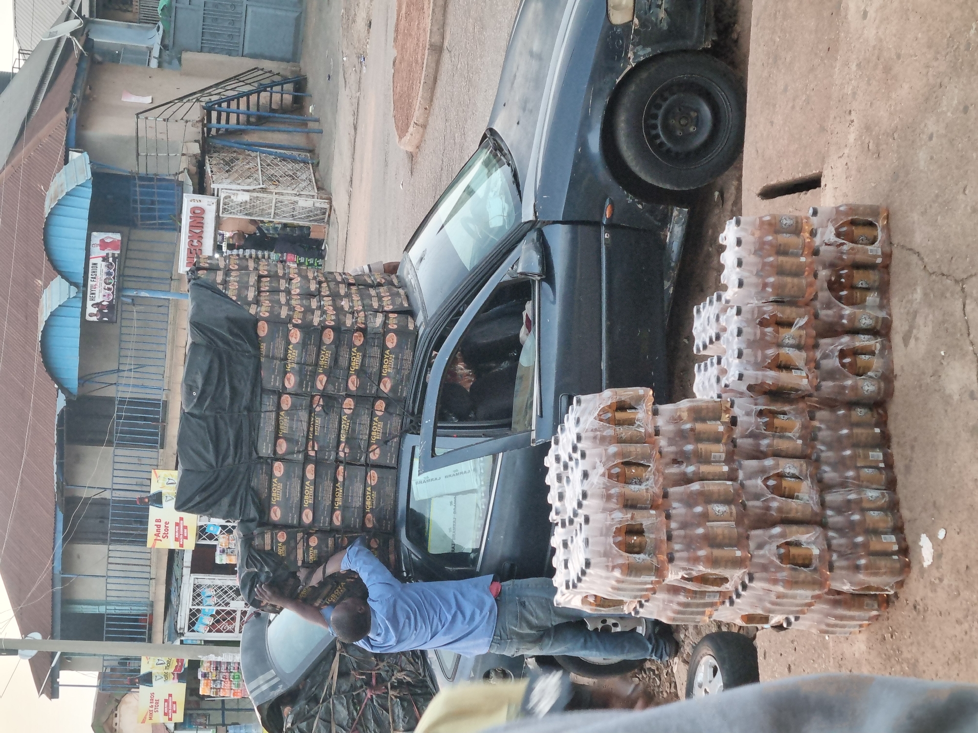 The image depicts a person loading a black car with cases of "Igoboya Bitters" beverages. The car is heavily loaded, with cases stacked high on the roof and alongside the vehicle. The setting is an urban street, with several stores visible in the background, including "Mike & Bros Store," "And B Store," "Henyul Fashion," and "Neckino." The scene appears to be in Jos, Nigeria, based on the visible signs and brands. The primary activity is the loading of the car with beverage cases.