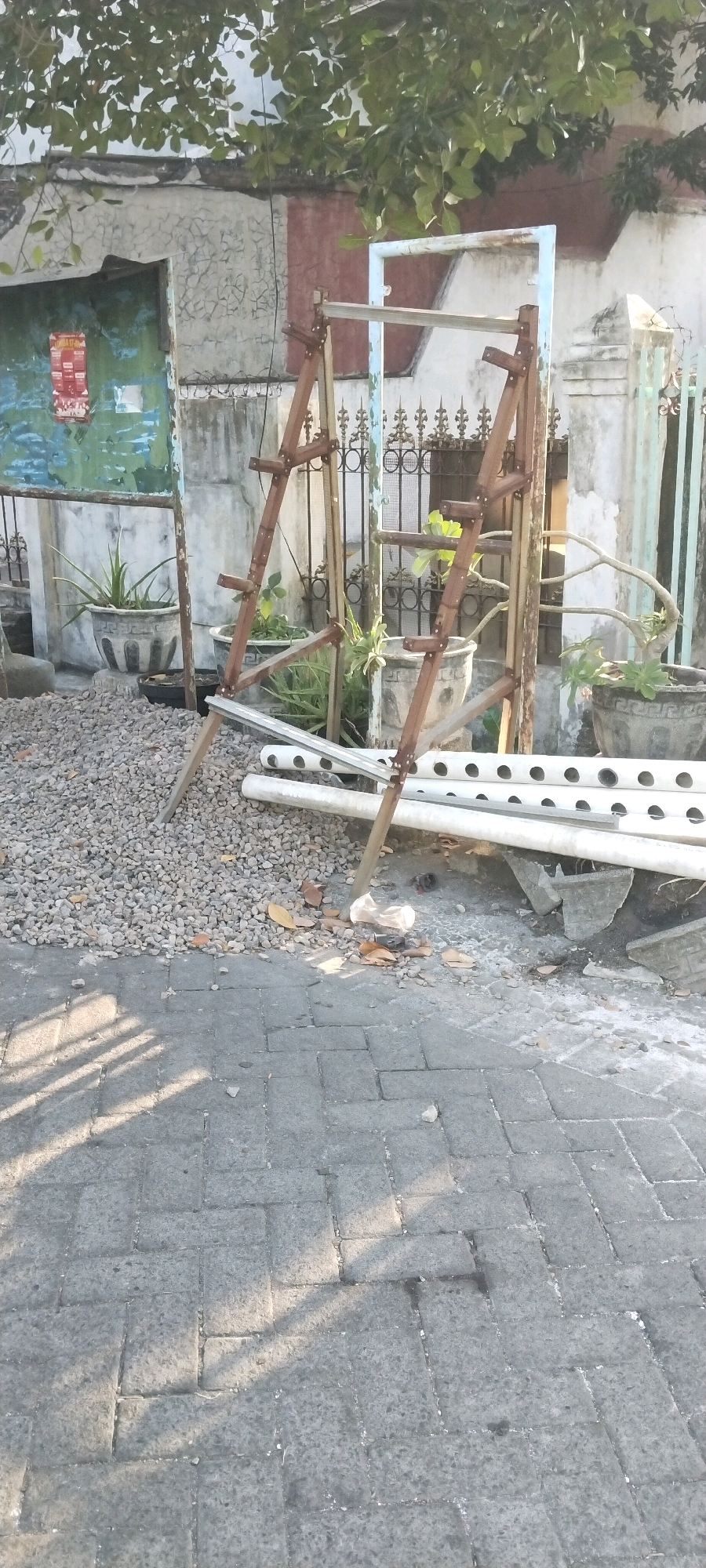 An outdoor scene in Surabaya, Indonesia, shows a narrow street or pathway paved with interlocking gray concrete blocks. The sunlight casts long shadows across the paving stones. To the left, a rough-textured, pale green wall with areas of cracked paint is visible, possibly part of a building. A dark green metal door or panel is set into this wall, with a red and white poster attached. In front of the wall, a decorative gray planter contains a spiky green plant.
Further into the scene, a rustic structure made of weathered wooden poles and crossbeams stands, supporting what appears to be a framework for a hydroponic system or similar setup. These wooden supports are assembled in an A-frame configuration. Lying on the ground nearby are several white PVC pipes with circular holes, suggesting they are part of a water channeling system. The ground around these pipes is covered with small, gray gravel.
Behind the wooden structure, a decorative metal fence with ornate scrollwork and vertical bars separates the immediate area from a building with a pale facade and some green metal bars or fencing. Several more gray planters, some with elaborate carved patterns, are present, containing various green plants. One planter in the background holds a small, gnarled tree with delicate branches. The overall impression is of an outdoor utilitarian or gardening area with some decorative elements.