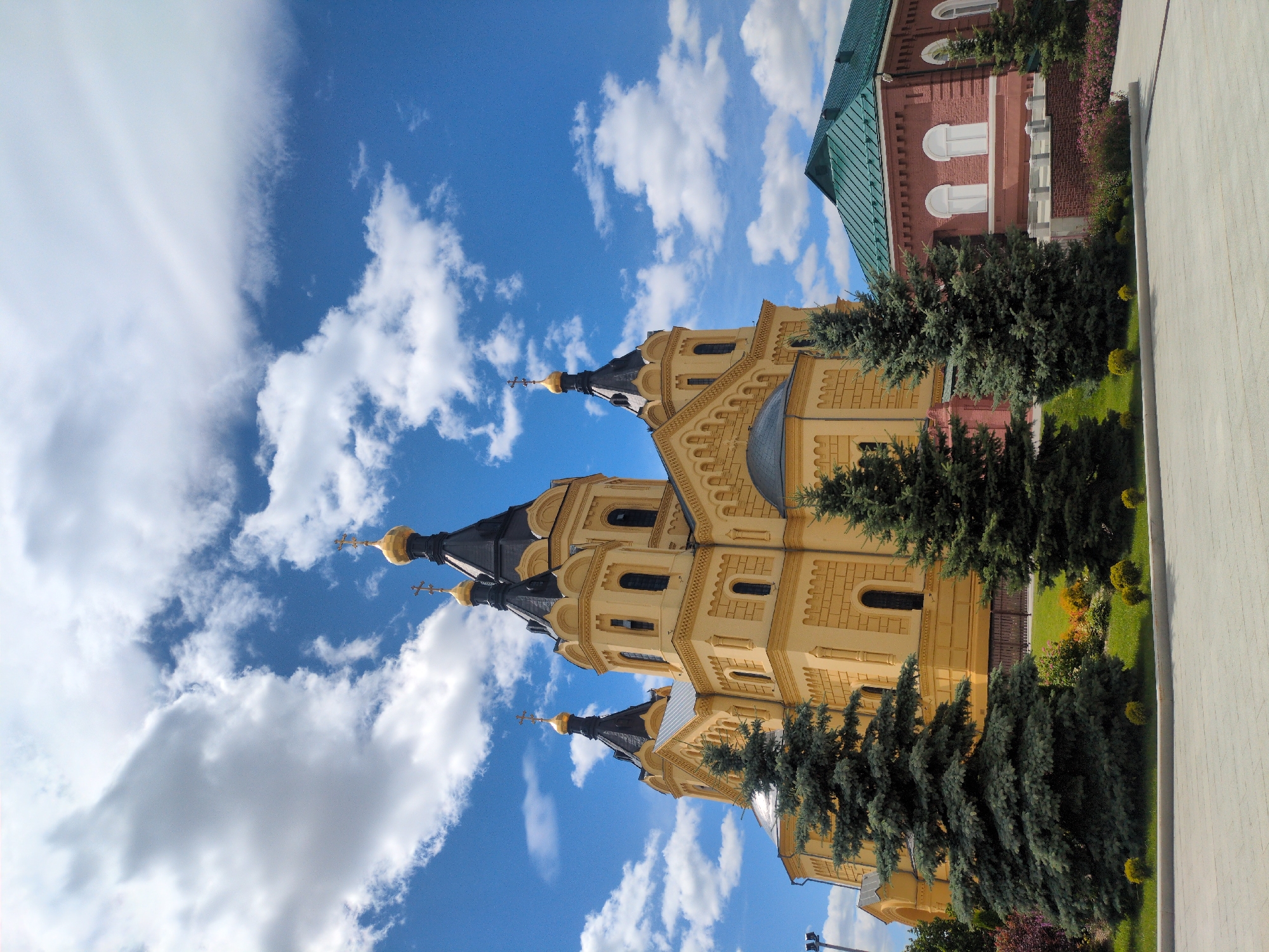The image depicts the Alexander Nevsky Cathedral in Nizhny Novgorod, Russia, under a bright blue sky with scattered white clouds. The cathedral, painted a warm yellow, features multiple onion domes topped with golden crosses, and dark gray roofs. To the right of the cathedral, a red brick building with a green roof and arched windows is partially visible. In the foreground, several tall evergreen trees stand on a well-maintained lawn, with a paved walkway in front of them. The scene is serene, with no people or animals present. The lighting suggests it is daytime, likely late morning or early afternoon, with sunlight illuminating the structures and foliage.
