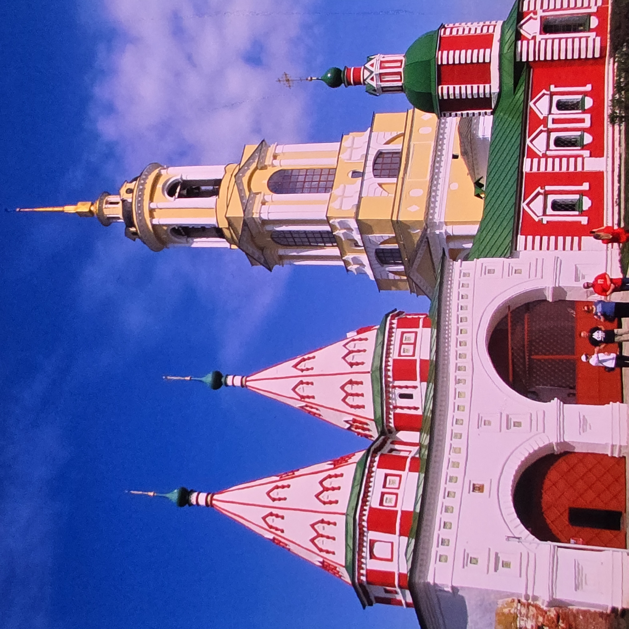The image depicts the Assumption Cathedral in Vladimir, Russia, under a clear blue sky. The cathedral features a prominent yellow bell tower with a gold spire, adorned with white trim and arched openings containing bells. Adjacent to the bell tower are two white conical roofs with red and white checkerboard patterns, topped with green onion domes. A separate red building with a green roof and white trim, featuring a green onion dome with a cross, is also visible. The foreground shows a white arched gate with a large red double door. Several individuals are present near the gate: one person in a white shirt and cap, another in a black t-shirt and khaki pants, a third in a blue top and dark pants, and a fourth in a red t-shirt and dark pants. Another person in a red dress is standing to the right. The ground is covered with grass.