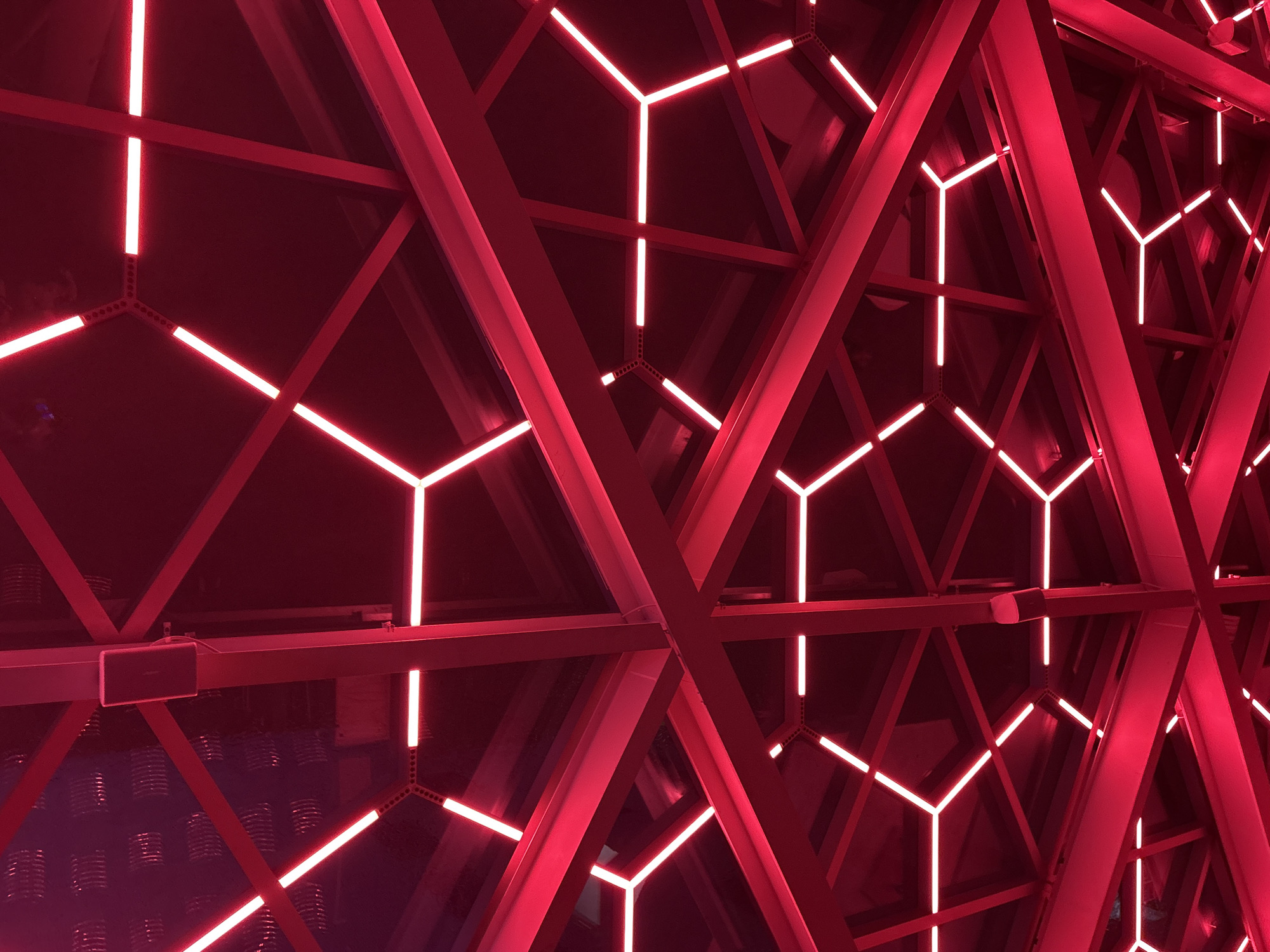 A low-angle shot captures a honeycomb-like structure with metallic supports and vibrant red neon lights forming hexagonal patterns. The structure is located in Mexico City. A speaker is mounted on one of the supporting beams. The overall lighting is low, adding depth and contrast to the scene.