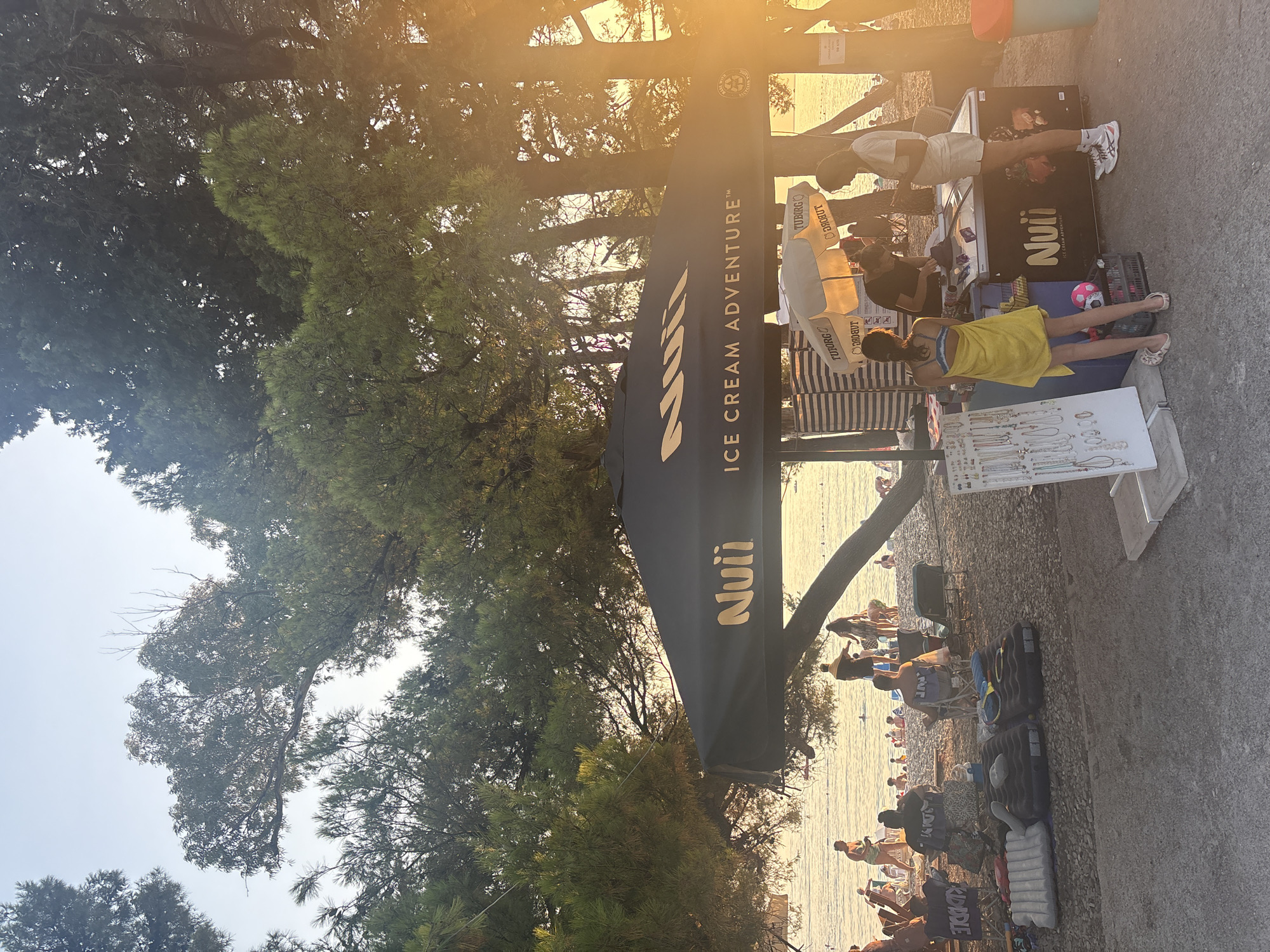 This is a medium-shot, eye-level, outdoor photograph taken at sunset on a beach in Bar, Montenegro. The foreground features a dark umbrella with "Nuii ICE CREAM ADVENTURE" written in gold letters, and a display stand with jewelry. Behind it, a beach scene unfolds with people sunbathing and swimming in the sea. To the right, a trash can is visible. The overall scene is bathed in the warm glow of the setting sun filtering through the trees.