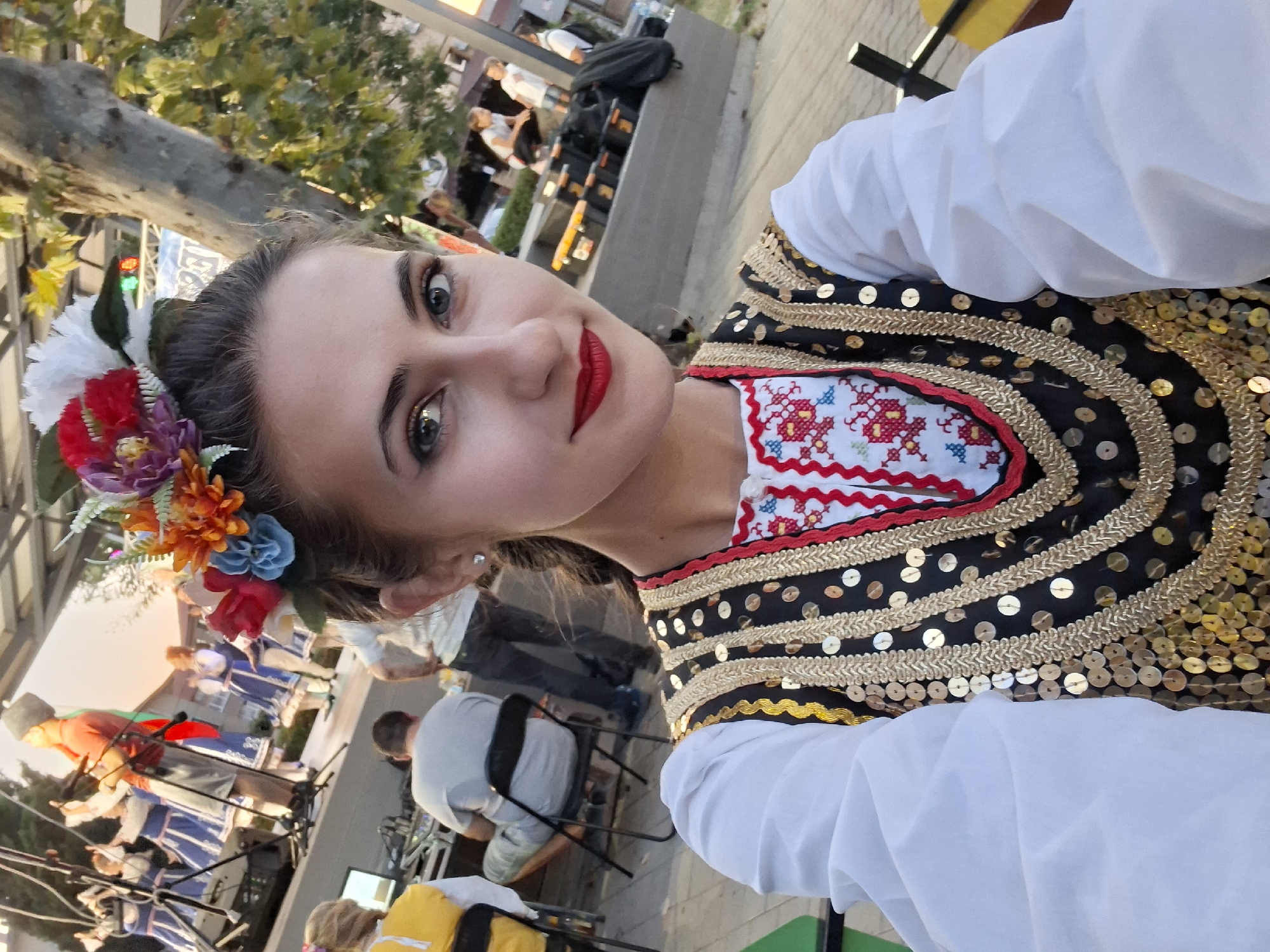 A young woman with a floral headdress and red lipstick poses for a selfie in a public outdoor setting, likely during a festival or cultural event. She wears a white blouse with voluminous sleeves and a decorative vest adorned with gold trim and sequins, over an embroidered garment. In the background, a group of people in traditional attire are performing on a stage, while others are seated or standing, observing the event. The environment appears to be a paved plaza with trees and an elevated stage, suggesting an urban or town square.