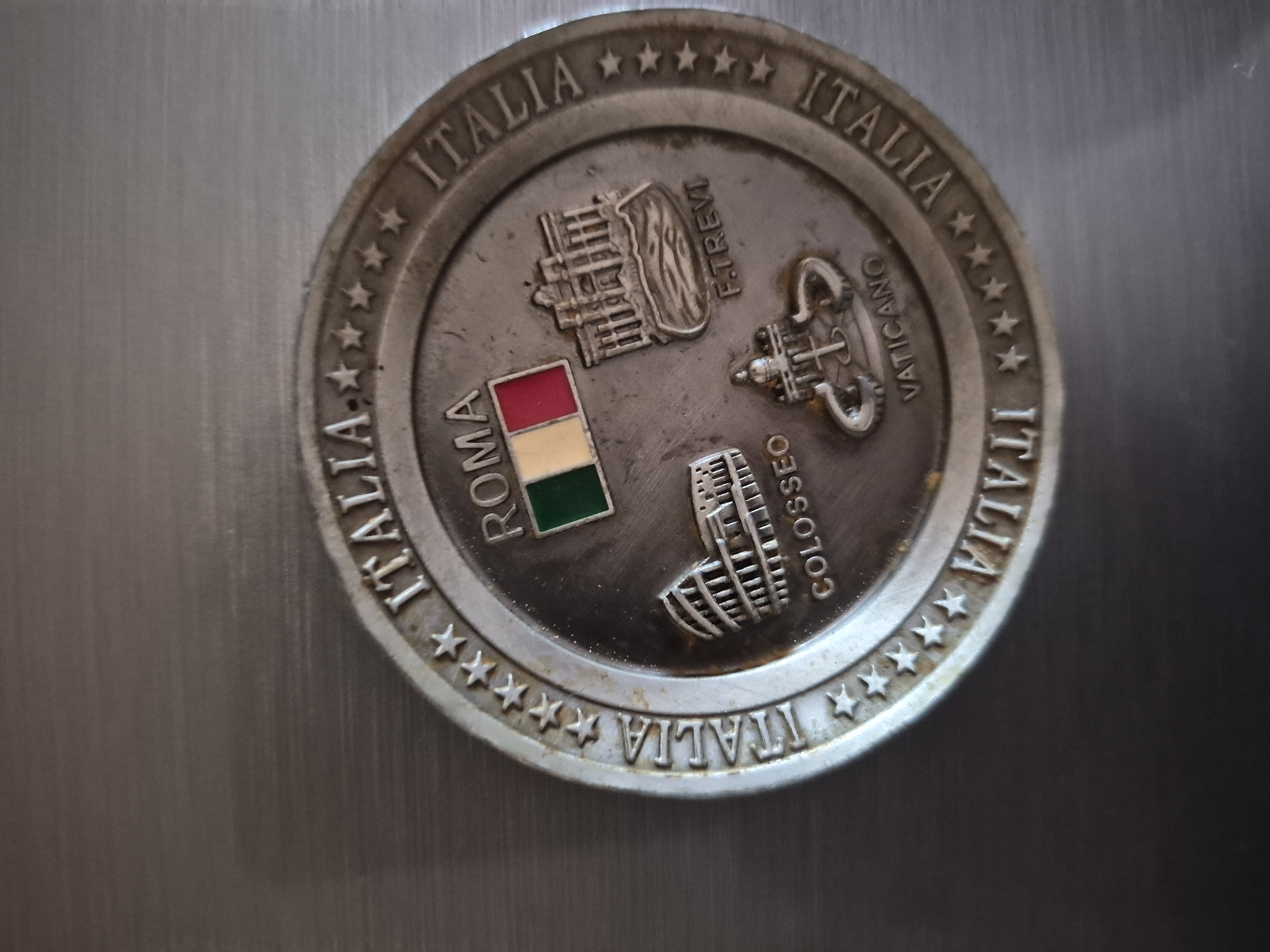 A circular, metallic souvenir coin or medal is depicted, resting on a brushed silver-toned surface. The coin has a slightly aged, silver-grey finish with visible patina and wear.
The central part of the coin features several embossed images and corresponding text, representing iconic Italian landmarks and symbols. A small, rectangular enamel inlay displays the Italian flag with its green, white, and red vertical stripes, situated next to the embossed word "ROMA." Above the flag, a detailed depiction of the Trevi Fountain is visible, labeled with "F. TREVI." To the right, symbols associated with the Vatican – a papal tiara and the Keys of Saint Peter – are embossed, accompanied by the word "VATICANO." Below these, the Colosseum is clearly rendered, with the text "COLOSSEO" placed beside it.
The outer rim of the coin features the word "ITALIA" repeated several times, separated by sequences of five-pointed stars. The entire composition highlights key historical and cultural aspects of Italy and its capital, Rome, including Vatican City. The background is a uniform, brushed metal surface, possibly a refrigerator door or a metal tabletop, reflecting minimal light. The lighting is soft and even, suggesting an indoor setting. There is no activity taking place, as it is a still image of an object.