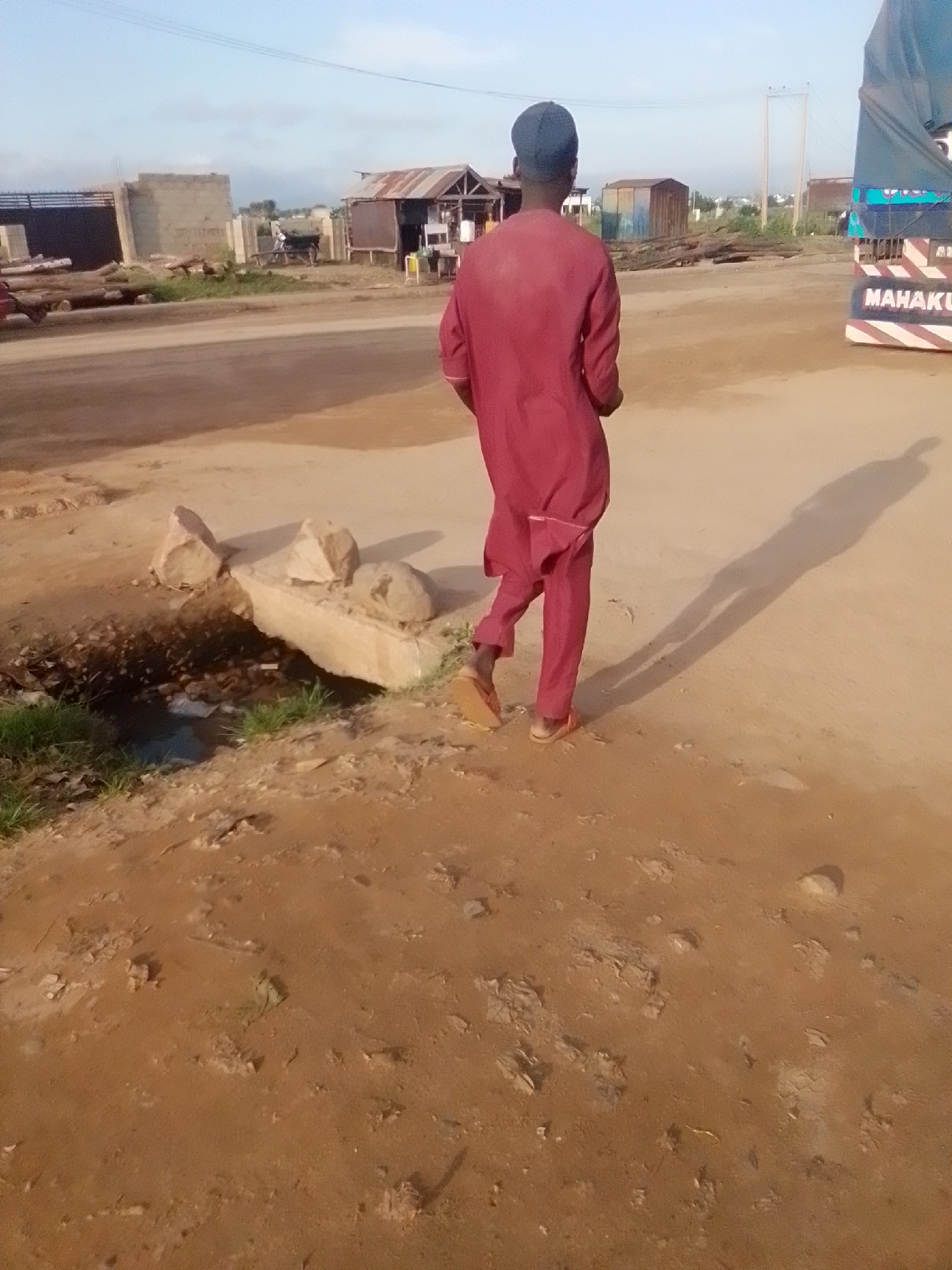 A person wearing a maroon traditional outfit and a dark cap walks away from the camera on a dusty, unpaved road in Kwadon, Nigeria. The person is casting a long shadow on the ground. In the background, there are several structures, including what appears to be a market stall with a corrugated metal roof, some unfinished buildings, and a large truck with the word "MAHAKU" visible on its side. The environment is dry and dusty, with scattered debris on the roadside. There are also some rocks and a small puddle of water near the foreground. The sky is clear with a few scattered clouds.