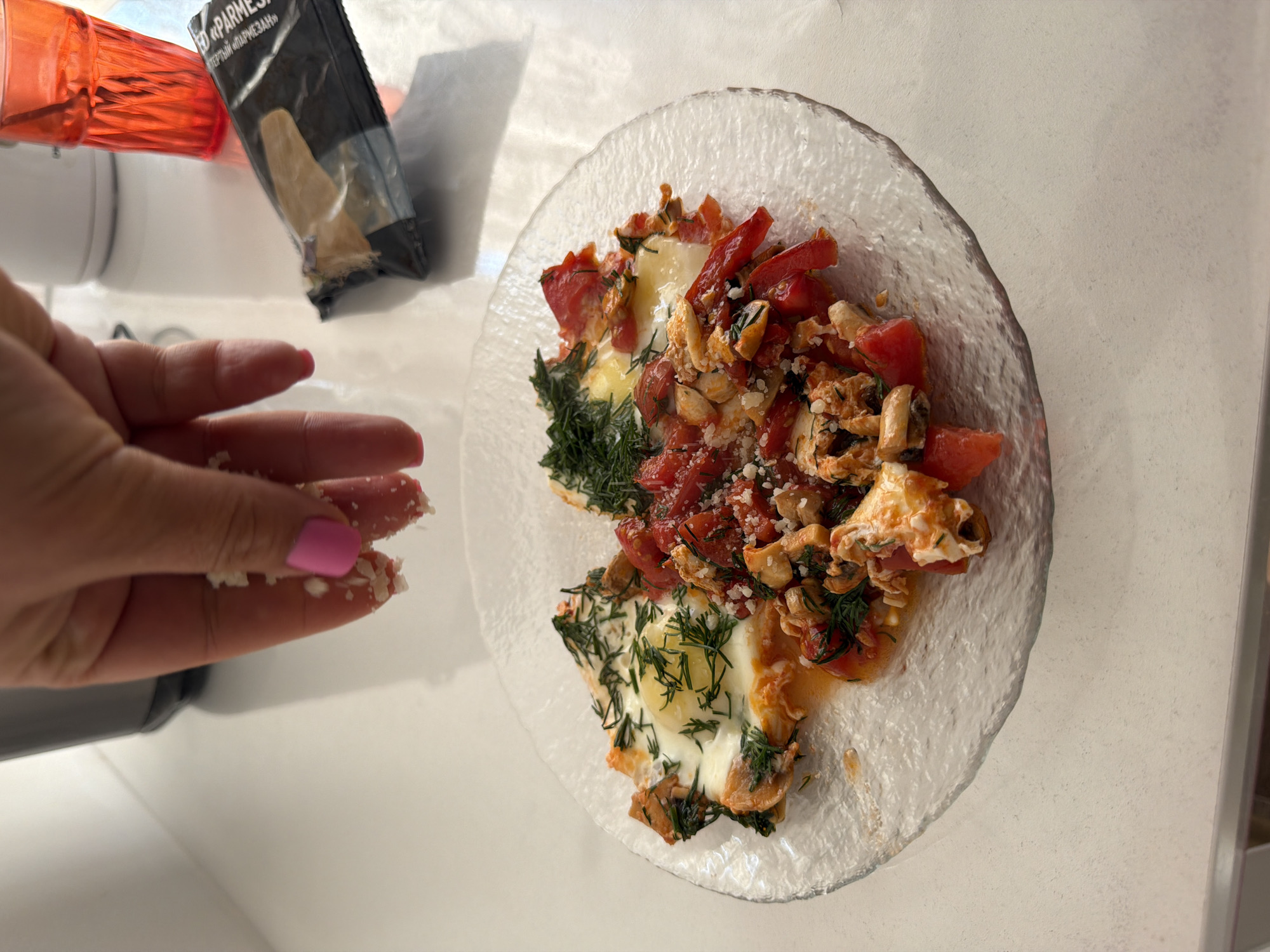 A close-up shot depicts a plate of food consisting of two fried eggs, diced tomatoes, red bell peppers, mushrooms, and dill, topped with grated cheese, being seasoned with cheese crumbles from a hand with pink fingernails. Next to the plate is a package labeled "Parmezan" in Russian. The location is presumably Leningradskaya, Russia, based on the "Parmezan" packaging. The scene is on a white countertop.