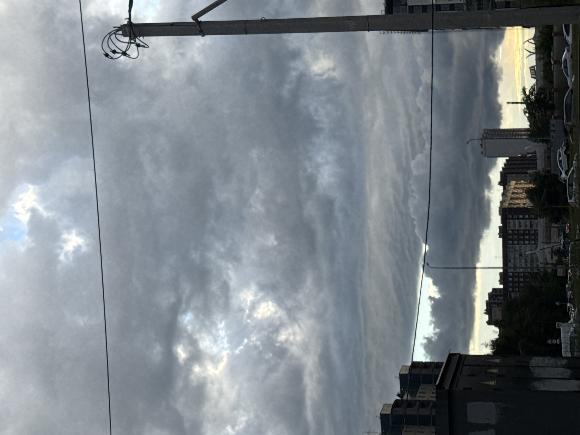 The image captures a view of a street scene in Saint Petersburg, Russia, under a dramatic sky. The perspective is low, looking upwards towards the clouds, which dominate the frame. These clouds are heavy and gray, with streaks of lighter clouds suggesting movement or wind. A portion of a utility pole with wires is visible in the foreground, adding a man-made element to the natural sky.
In the background, the silhouette of a city skyline emerges. Several multi-story buildings are visible, some appearing to be residential blocks with numerous windows. The architecture suggests a modern urban environment. There are also hints of trees and greenery interspersed among the buildings, softening the urban landscape. On the right side of the image, a street with parked cars and a street lamp can be seen, indicating a populated area.
The light in the sky suggests it is either early morning or late evening. The horizon is lit with a soft, warm glow, contrasting with the darker, overcast clouds overhead. This creates a sense of atmospheric depth and perhaps a hint of an approaching or receding storm. The overall mood is somewhat somber, given the overcast sky, but the light on the horizon offers a touch of hope or transition. There is no visible text in the image.