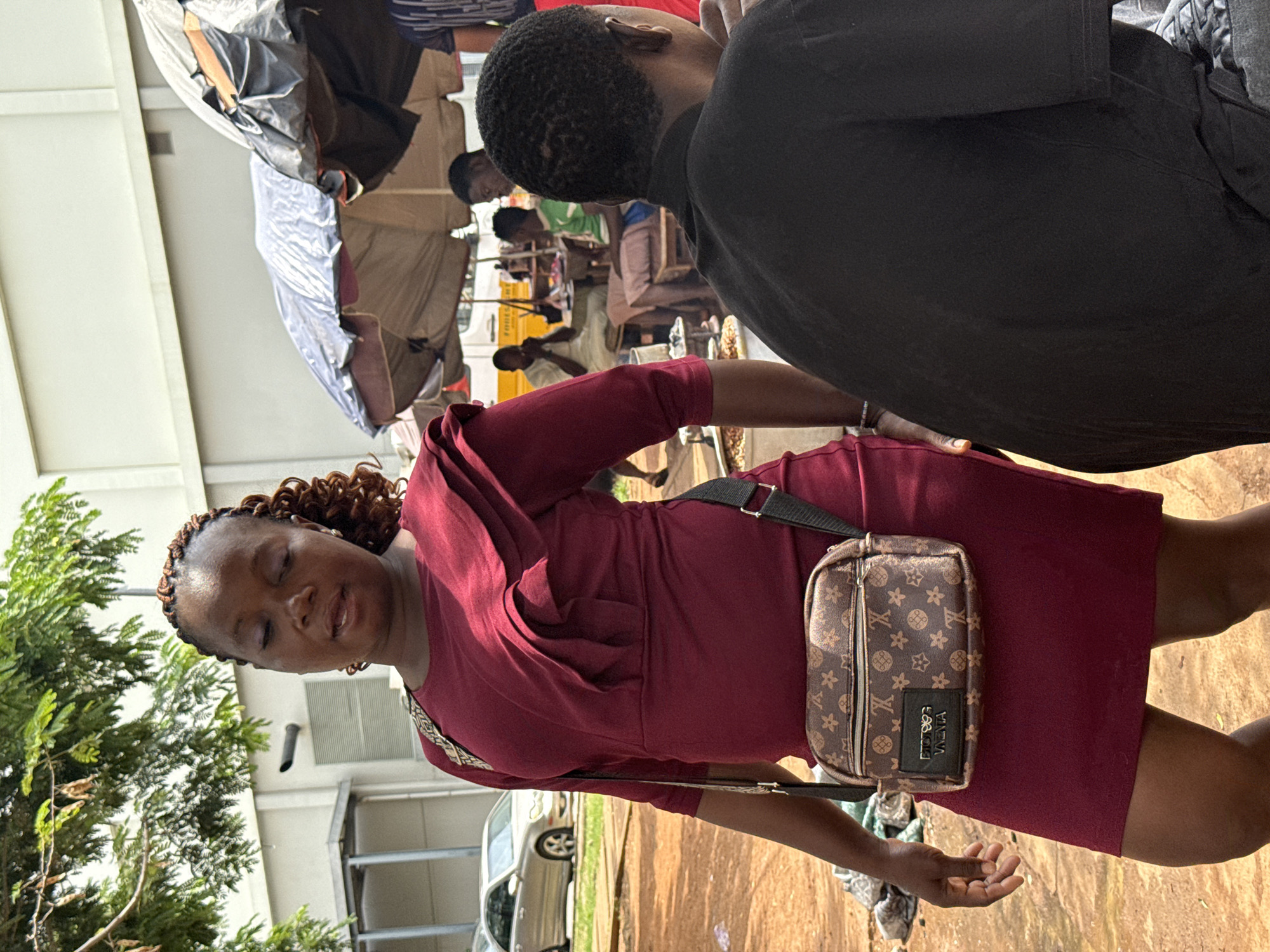 The image shows a woman in a maroon dress with a patterned crossbody bag. She has braided hair and is looking downwards with her eyes closed. In the background, there are several other people, some sitting at tables, suggesting a market or outdoor gathering. There are also some tents or awnings visible, indicating a potentially sunny day in an outdoor setting. A silver car is parked in the background, and a tree with green foliage is to the left of the woman. The overall scene appears to be an informal, bustling environment. The woman's expression could suggest contemplation or relaxation. No visible text is present in the image.