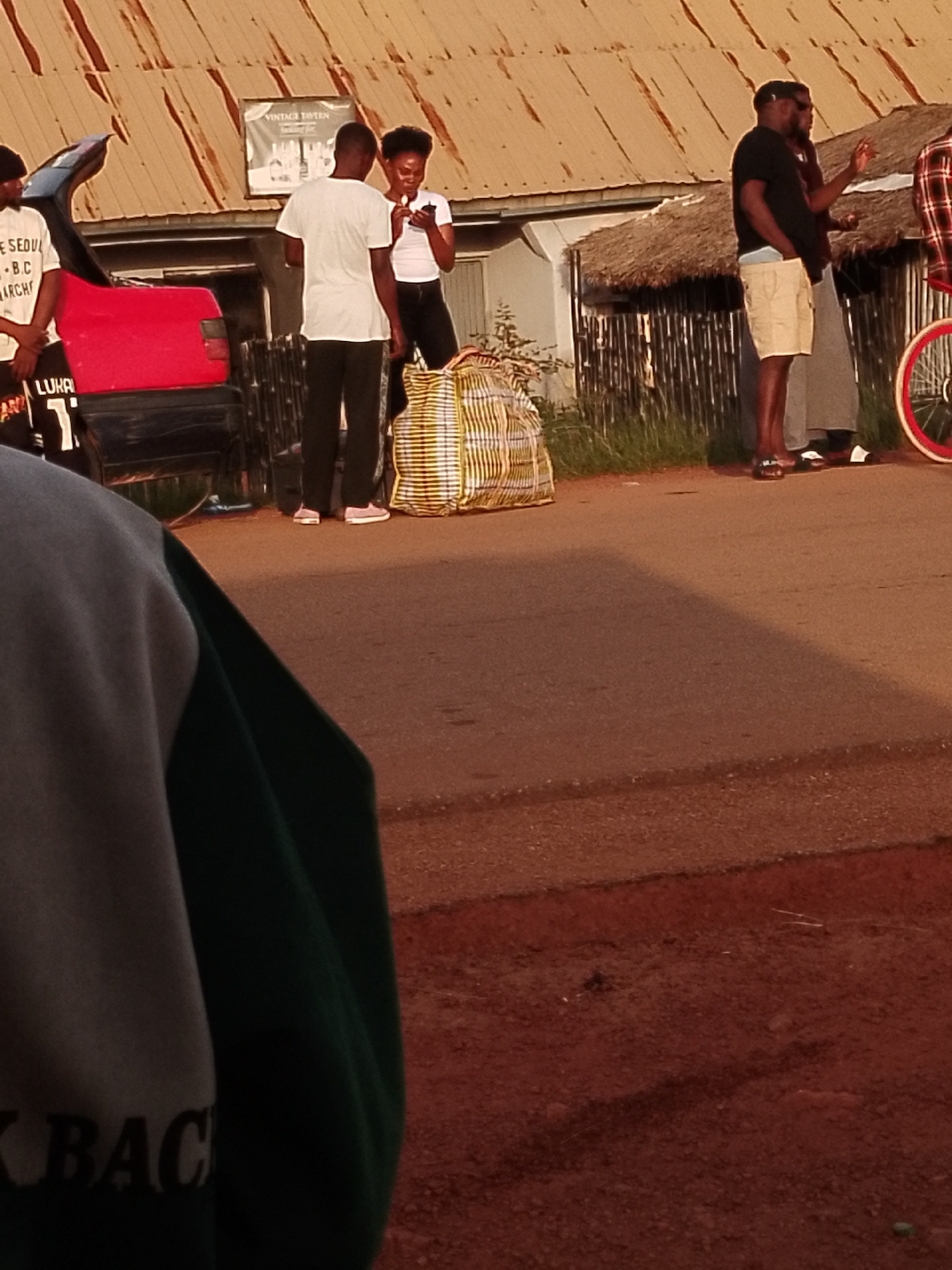 The image depicts a street scene in Agwala, Nigeria, with several individuals gathered around a vehicle and a building. In the foreground, a person is partially visible, wearing a grey and green garment with the word "BACK" printed on it. To the left, a red vehicle's trunk is open, and a person in a white t-shirt with text is standing beside it.
In the middle ground, two men and a woman are interacting. One man wears a white t-shirt and dark pants, while the other wears a black t-shirt and light-colored shorts. The woman is in a white t-shirt and dark pants, holding a mobile phone. Behind them, a large, striped bag is placed on the ground. The building in the background features a corrugated iron roof and a thatched section. A sign above a doorway reads "VINTAGE TAVERN."
Further to the right, another individual is seen wearing a plaid shirt, with a bicycle with red rims visible. The ground is paved with asphalt, and a dirt path is present on the right side of the image. The overall lighting suggests it is either early morning or late afternoon.