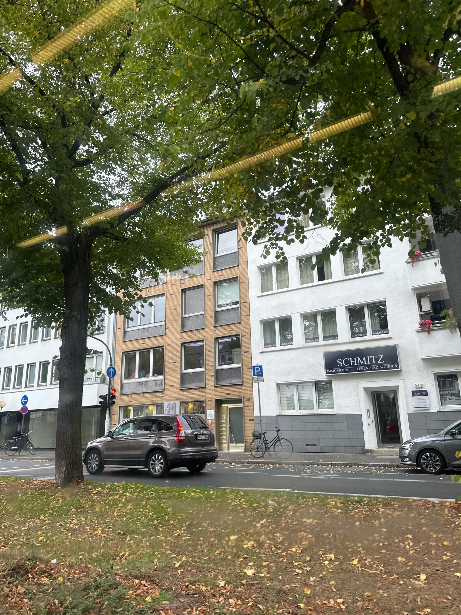 The image shows a street scene in Krefeld, Germany, viewed from a slightly elevated perspective, possibly from a vehicle. The foreground consists of a grassy embankment covered with fallen yellow and brown leaves, indicating an autumnal season. Beyond the embankment is a street with a single lane of traffic. A brown SUV is stopped at a red traffic light.
To the right of the street are several multi-story buildings. The closest building on the right is white with several windows and balconies adorned with pink flowers. A prominent black sign above the entrance reads "SCHMITZ IMMOBILIEN // LEBEN UND WOHNEN". Further to the left, there is a building with a yellow brick facade and more windows. To the left of that, a white building with rows of windows is visible.
A large tree with green foliage stands on the left side of the image, partially obscuring the buildings. A street sign with a blue arrow pointing left is visible near the traffic light. A bicycle is parked on the sidewalk in front of one of the buildings. The sky is overcast. The scene is static, with no visible human activity beyond what can be inferred from the presence of the SUV.