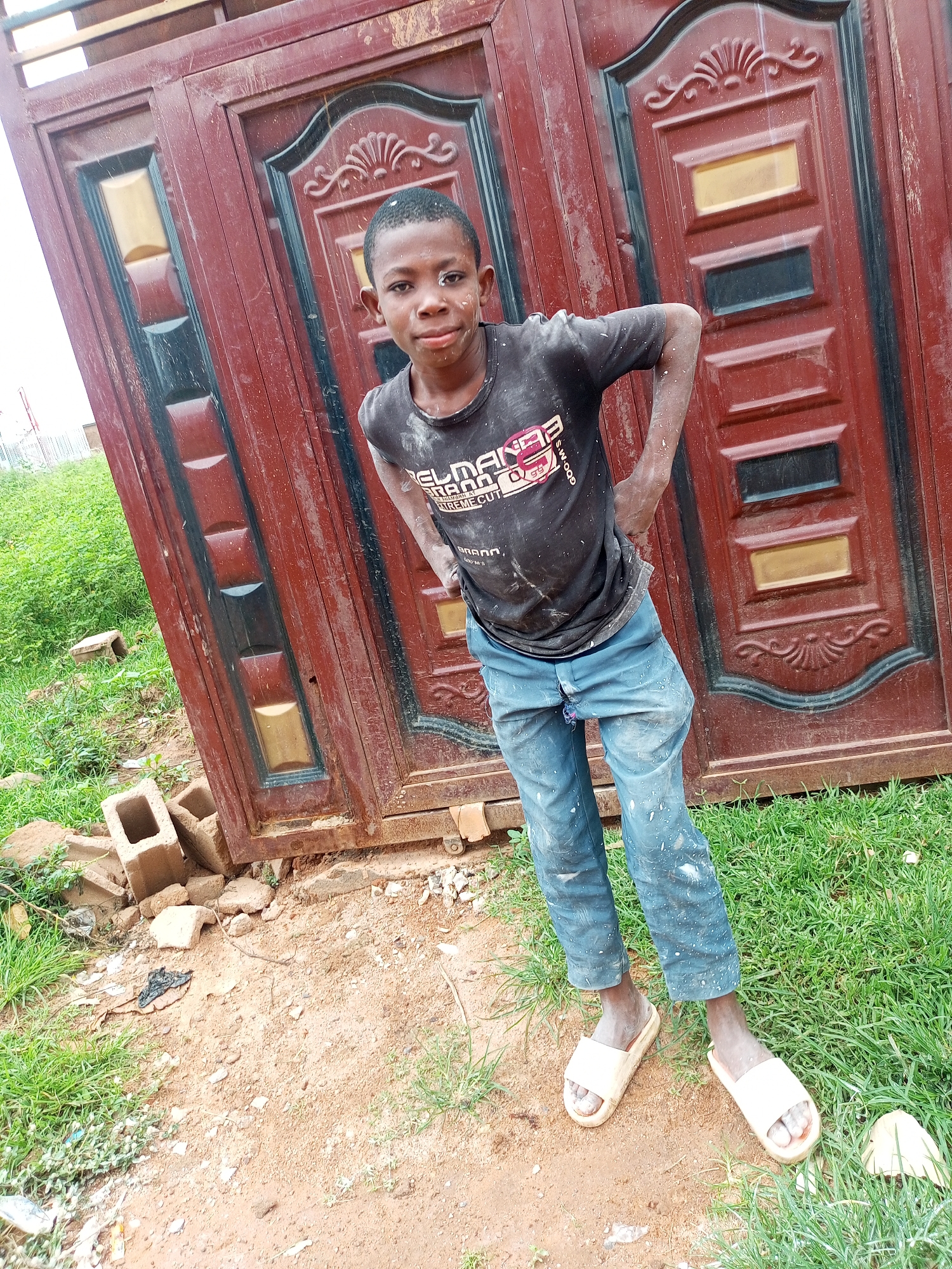 A young boy with dust on his face and body stands in front of a dark red, ornate double door. He is wearing a black t-shirt with white lettering and light blue pants that are also dusted. He has white sandals on his feet. The ground in front of the door is a mix of dirt and grass, with some scattered bricks and debris. The background is a lush green grassy area. The scene appears to be outdoors, possibly in a residential or construction area in Tempure, Nigeria.