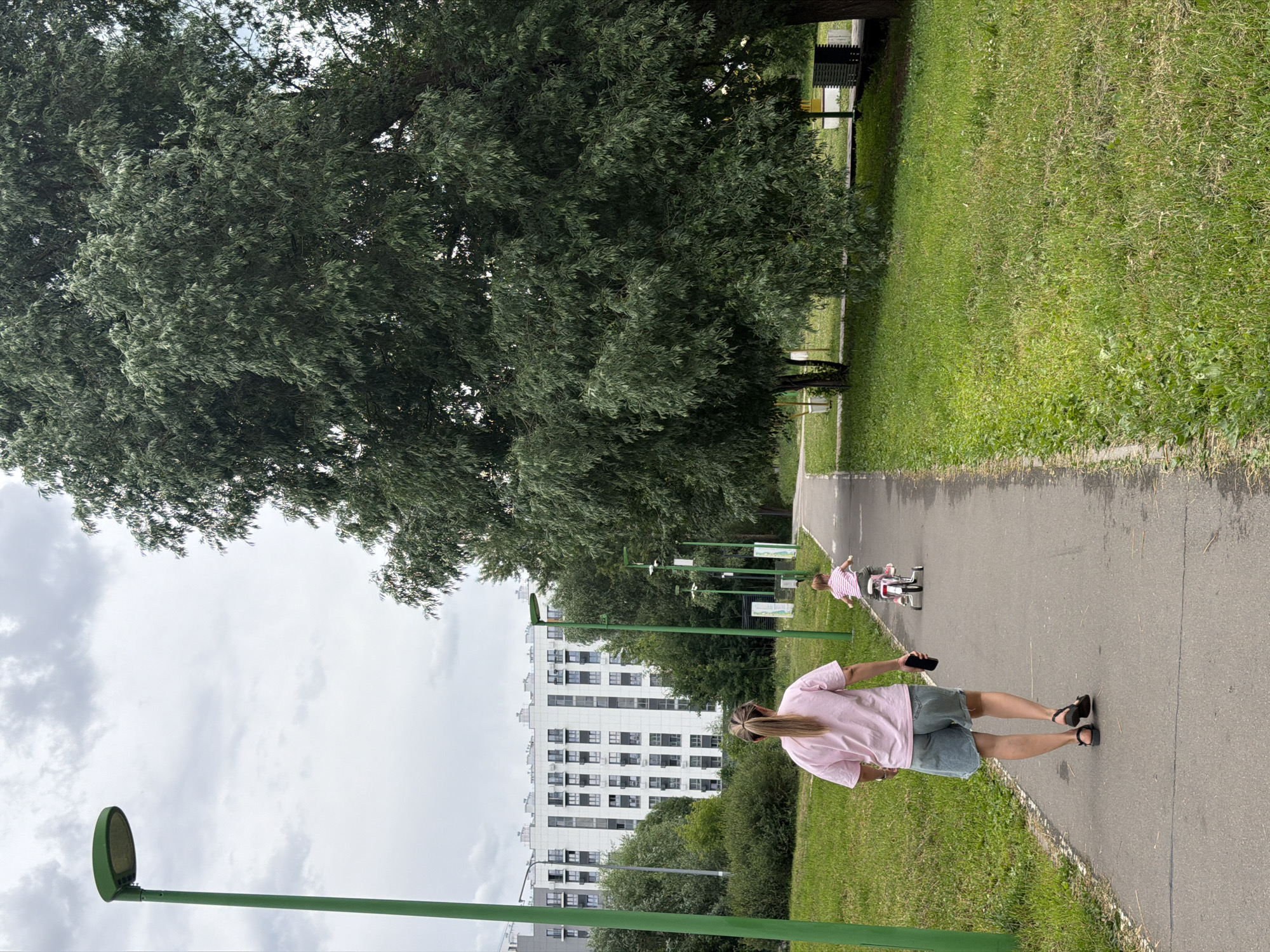 An outdoor scene depicts a paved pathway in a park-like setting with a large, leafy tree to the right and a white, multi-story building in the background. A woman wearing a pink t-shirt and denim shorts walks away from the camera on the pathway, holding a dark smartphone in her right hand. A young child on a pink tricycle is in front of her, also moving away from the camera. The pathway is bordered by green grass and a sidewalk. Several green lamp posts are visible along the pathway. The sky is overcast with gray clouds.