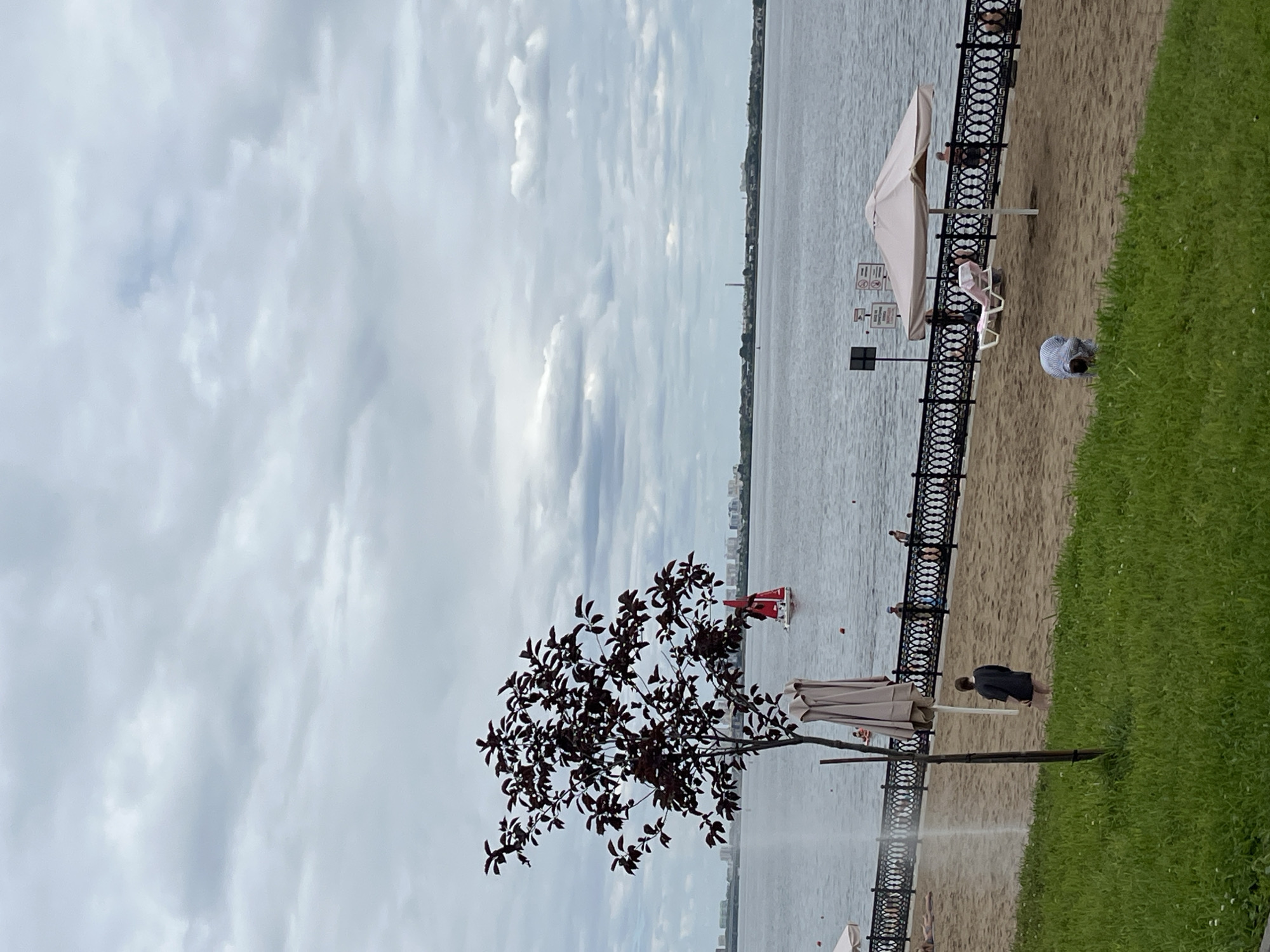 A wide shot captures a cloudy day at a city beach. The foreground shows lush green grass. The midground is a sandy beach with people relaxing and walking. A black metal fence separates the beach from the water. A small tree with dark leaves stands on the left side of the beach. In the water, a red sailboat is visible in the distance. The background features a cityscape with buildings. The overall scene suggests a calm, recreational day at a waterfront location.