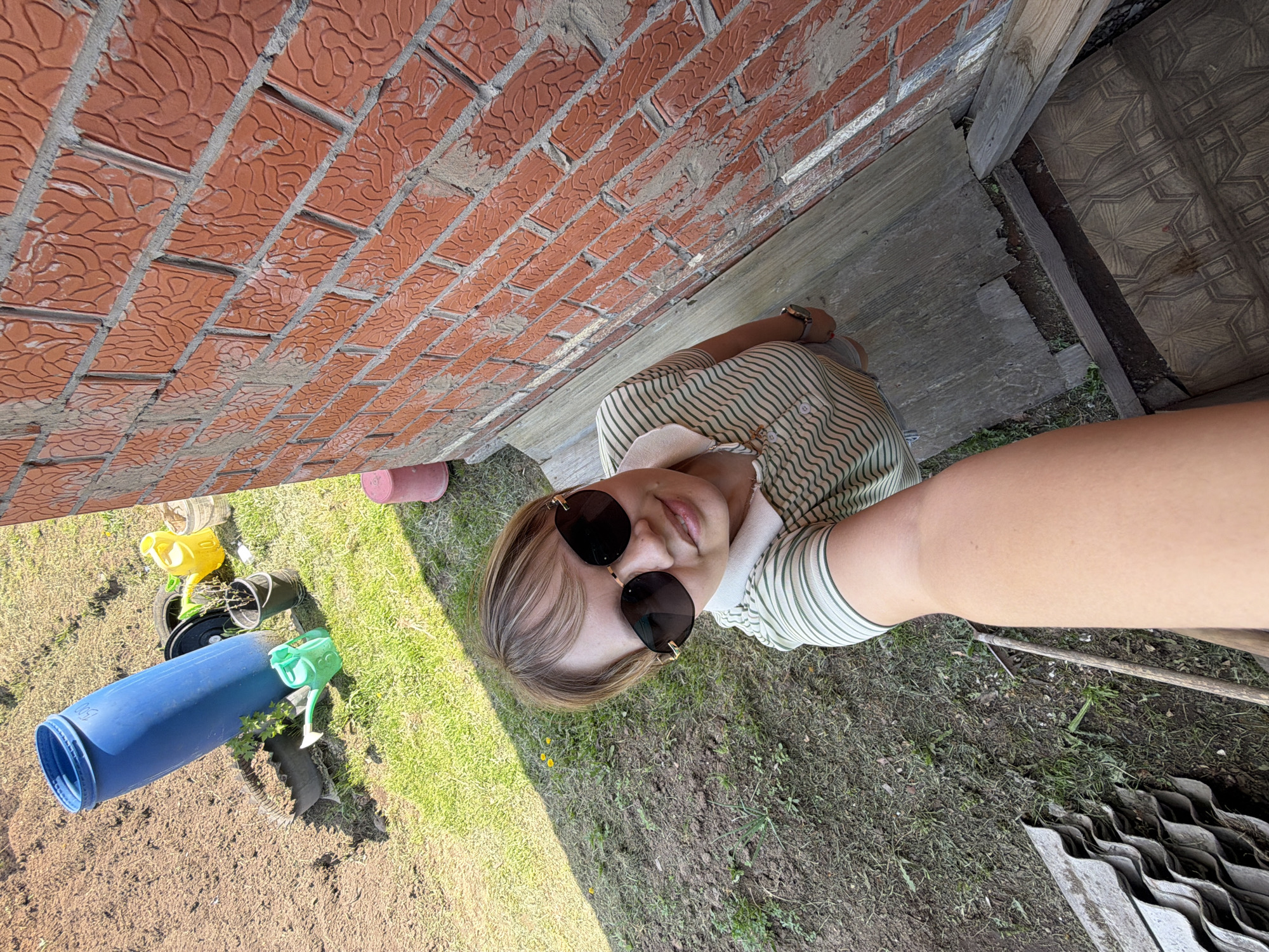 The image appears to be a selfie taken outdoors near a building. The subject is a person with blonde hair wearing sunglasses and a striped shirt. They are positioned in front of a brick wall and a wooden structure, possibly a house or shed. The ground is covered in grass and dirt, with gardening tools and containers scattered around. The presence of corrugated metal sheets suggests possible construction or renovation activities. There is no evidence to confirm the location as the city of Чумна, Russia.
