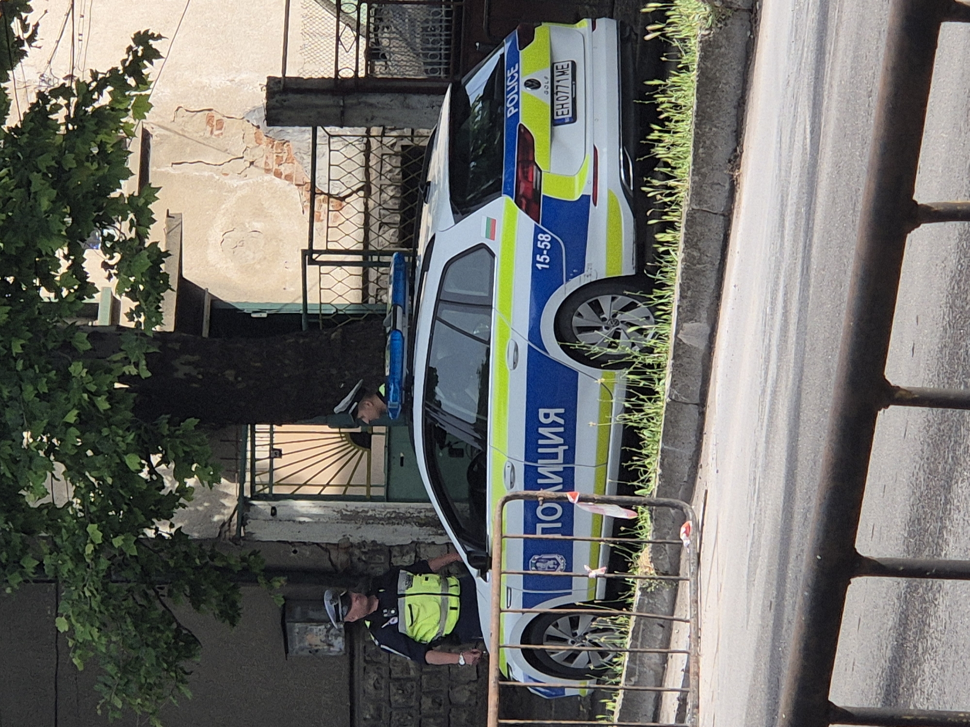 The image, rotated 90 degrees counter-clockwise, captures a daytime scene featuring a police car and two officers on a street in what appears to be Bulgaria.
**Subjects:** The primary subjects are a white Volkswagen Golf police car and two police officers. One officer, wearing a dark uniform, reflective neon yellow vest, and cap, stands next to the front door of the police car. The second officer, also in a dark uniform and cap, is partially visible further back, near the rear of the car, seemingly obscured by the car window and a background building. Surrounding elements include an old building, a green-leaved tree, a street, a grassy curb, and a metal barrier in the foreground.
**Activity:** The police car is parked on the side of an asphalt road. The officers appear to be on duty, stationed near their vehicle. There is no specific active event depicted, but rather a moment of police presence.
**Notable details:** The police car is distinctive with its white base, prominent blue and neon yellow stripes, and blue light bar on the roof. Visible text on the car includes "ПОЛИЦИЯ" (Police in Cyrillic) on the side, "POLICE" on the rear, the car model "GOLF," and the unit number "15-58." A small Bulgarian flag is also visible on the car's side, and the license plate reads "EH 0771 ME," which is a Bulgarian plate, further confirming the location. The background features an old building with a light-colored, somewhat dilapidated facade and a decorative metal gate or balcony. A lush green tree stands to the left, and a rusty metal fence acts as a foreground element on the right. The scene is well-lit, indicating clear, sunny weather.
**Visible text:** "ПОЛИЦИЯ", "POLICE", "GOLF", "15-58", and "EH 0771 ME".