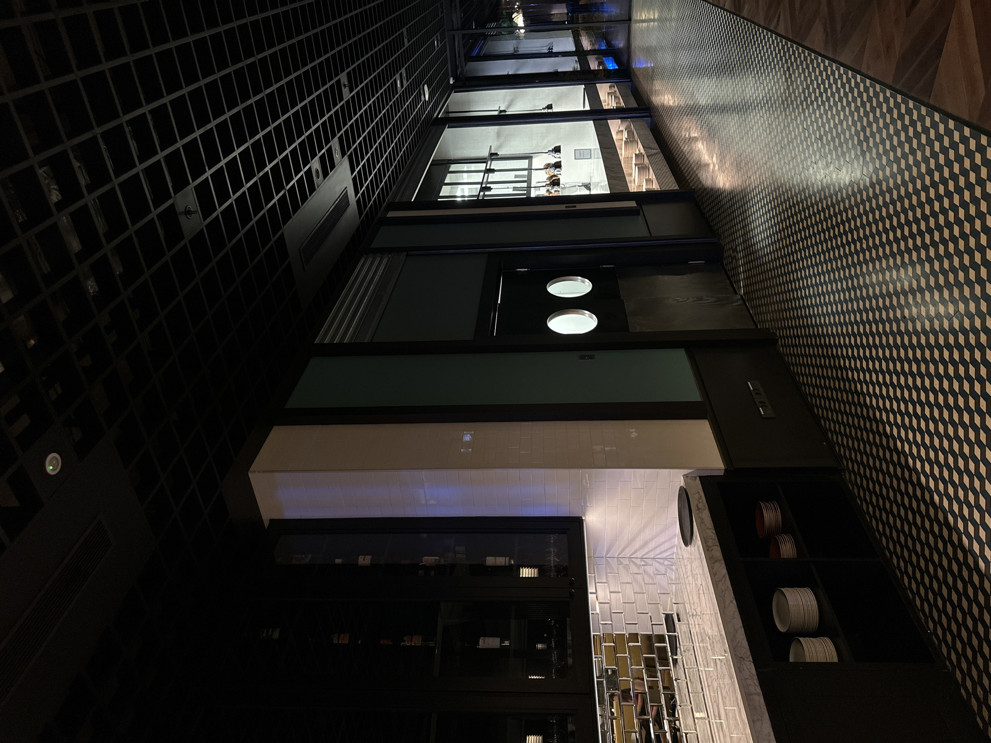 The image depicts the interior of a building or business, likely a restaurant, with a modern design. The walls are decorated with a repeating cube pattern in black and white, while a section of the wall features a display cabinet filled with wine bottles and other items. Above, there are two round windows or lights in the wall. The scene is illuminated with dim lighting, creating a dark and atmospheric ambiance. The location is not specified, nor are any specific details about Batumi, Georgia provided. No people or specific actions are visible in the image.