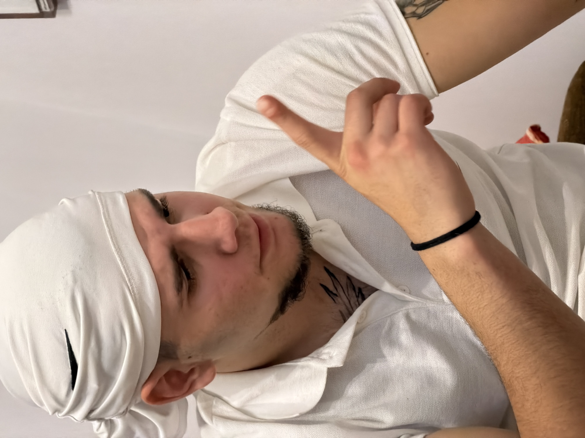 A man is shown wearing a white head covering and shirt. His eyes are closed, and he has a light beard. A tattoo is visible on his neck. He is making a hand gesture with his right hand. The location is not discernible from the image.