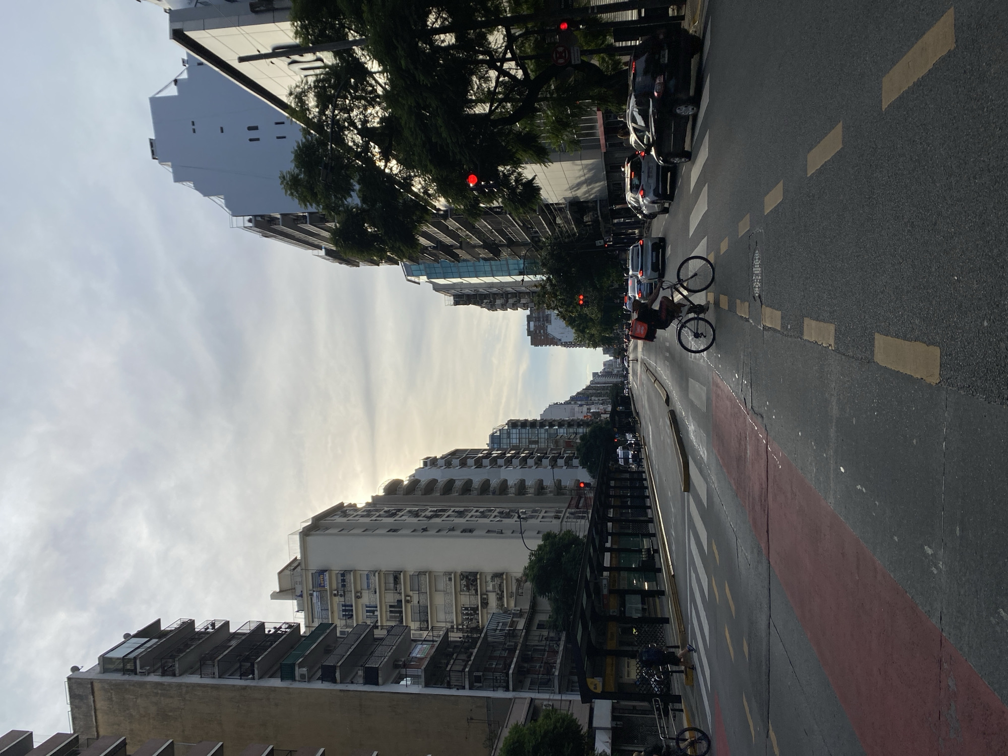 The image captures a street scene in Buenos Aires, Argentina. The perspective is looking down a wide avenue flanked by tall, predominantly white, apartment buildings with balconies. The street has multiple lanes of traffic, with cars visible on the right side. A bicycle rider, presumably a delivery person with a red bag, is visible mid-street near a pedestrian crosswalk marked with alternating white and red stripes. Streetlights are present. The sky is overcast with sun rays visible.