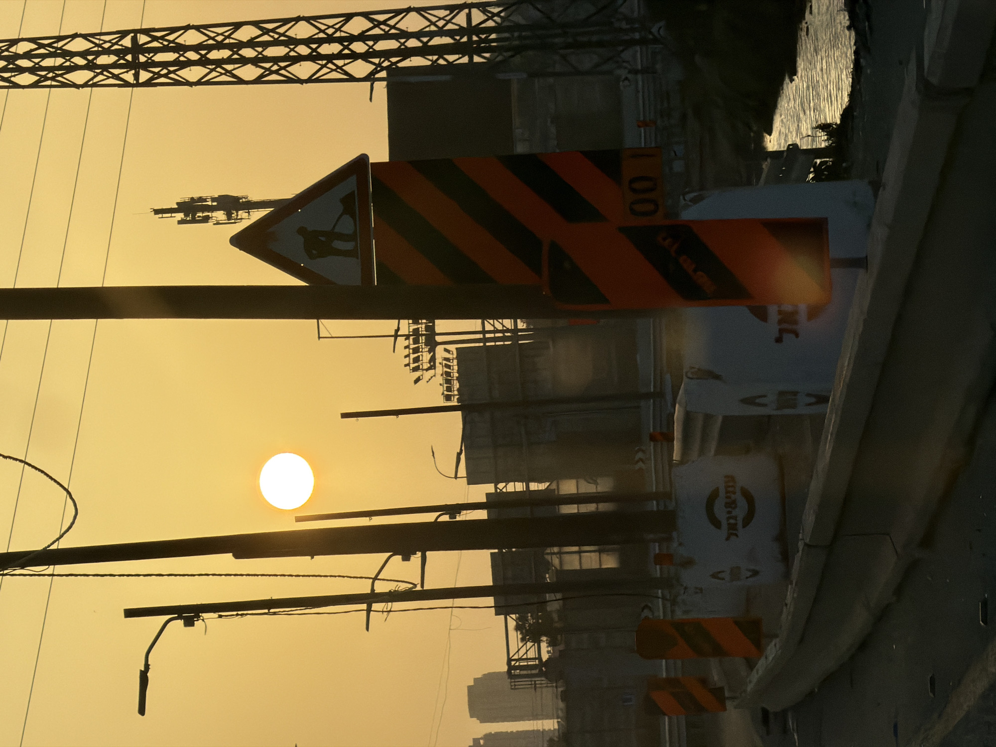 The image captures an urban or industrial landscape at either sunrise or sunset, characterized by warm golden and orange hues dominating the sky. The scene, located in Azor, Israel, features prominent silhouettes of structures against the bright sky.
In the upper portion of the frame, a large, intricate metal lattice structure, possibly part of a bridge or power transmission tower, stretches across. Below it, several thick, dark horizontal poles with attached wires and lighting fixtures traverse the sky. A bright, circular white sun is visible in the middle-left, casting long shadows and creating a striking contrast with the dark foreground elements.
The foreground and midground contain several construction or road-related objects. A triangular warning sign with a black silhouette of a person shoveling dirt is visible, indicating road work. Nearby, multiple rectangular signs with distinctive black and orange diagonal stripes are present, commonly used for construction zone warnings. One such sign partially reveals the numbers "00 1". On the right side, a series of white barriers or drums are aligned. Hebrew text is visible on some of these barriers; one clearly displays "פיבי בראל" (Pibi Barel), suggesting a construction company, and another partially shows "אל" (El).
In the background, dark silhouettes of buildings and other urban structures are visible, hinting at a developed area. To the far right, a body of water, possibly a river or canal, shimmers with the reflection of the low sun. A dark, elevated path or road runs alongside this water body. The overall impression is one of ongoing development or infrastructure within an urban setting, beautifully illuminated by the dawn or dusk light.