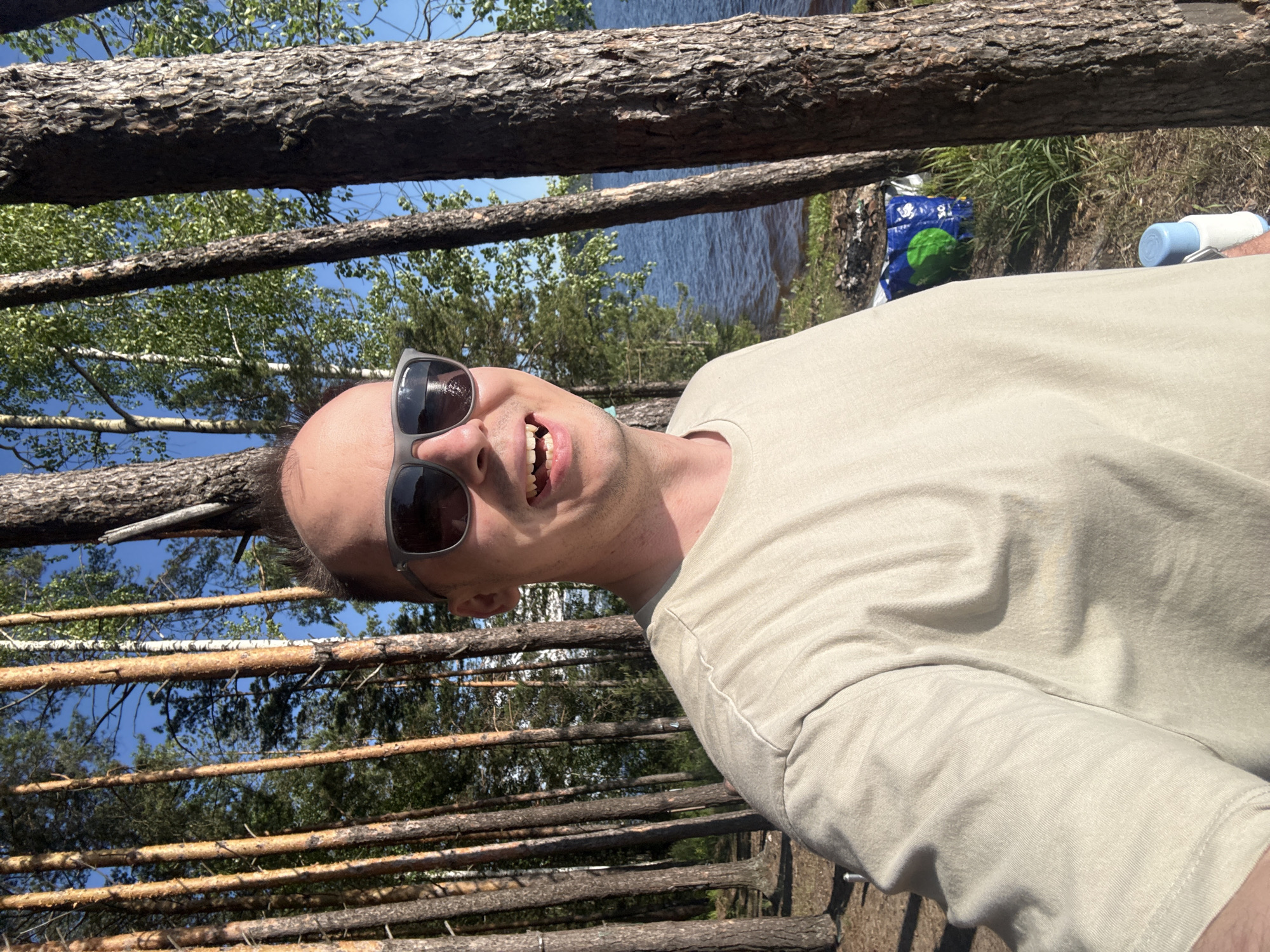 The image shows a man taking a selfie outdoors. He is wearing sunglasses and a light-colored t-shirt. Behind him, there are tall pine trees and a body of water, likely a lake or river. The sun is shining, casting shadows. In the background, a blue and green bag is visible on the ground, possibly for camping supplies. The setting appears to be a natural landscape in or near Gora (Khokhlovskoe S/P), Russia, likely a recreational area or campsite.