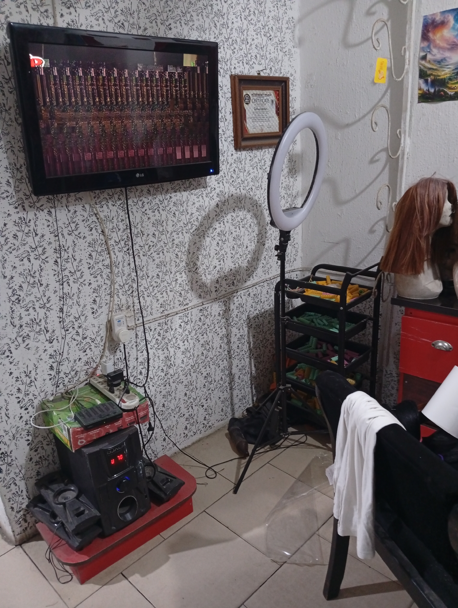 The image shows a room interior. A black LG television is mounted on a wall with a floral patterned wallpaper. Below the TV, a red side table holds a black speaker system and a green box with electronic devices. Various cords and plugs are visible on the wall. To the right of the TV, a framed certificate hangs on the wall. A ring light is positioned on a tripod next to a black rolling cart filled with yellow and green objects. A mannequin head with a brown wig sits on a red cabinet. A black chair with a white cloth draped over it is in the foreground. The floor is tiled. The location, Lokoja, Nigeria, is not deducible from the image. No people are visible.