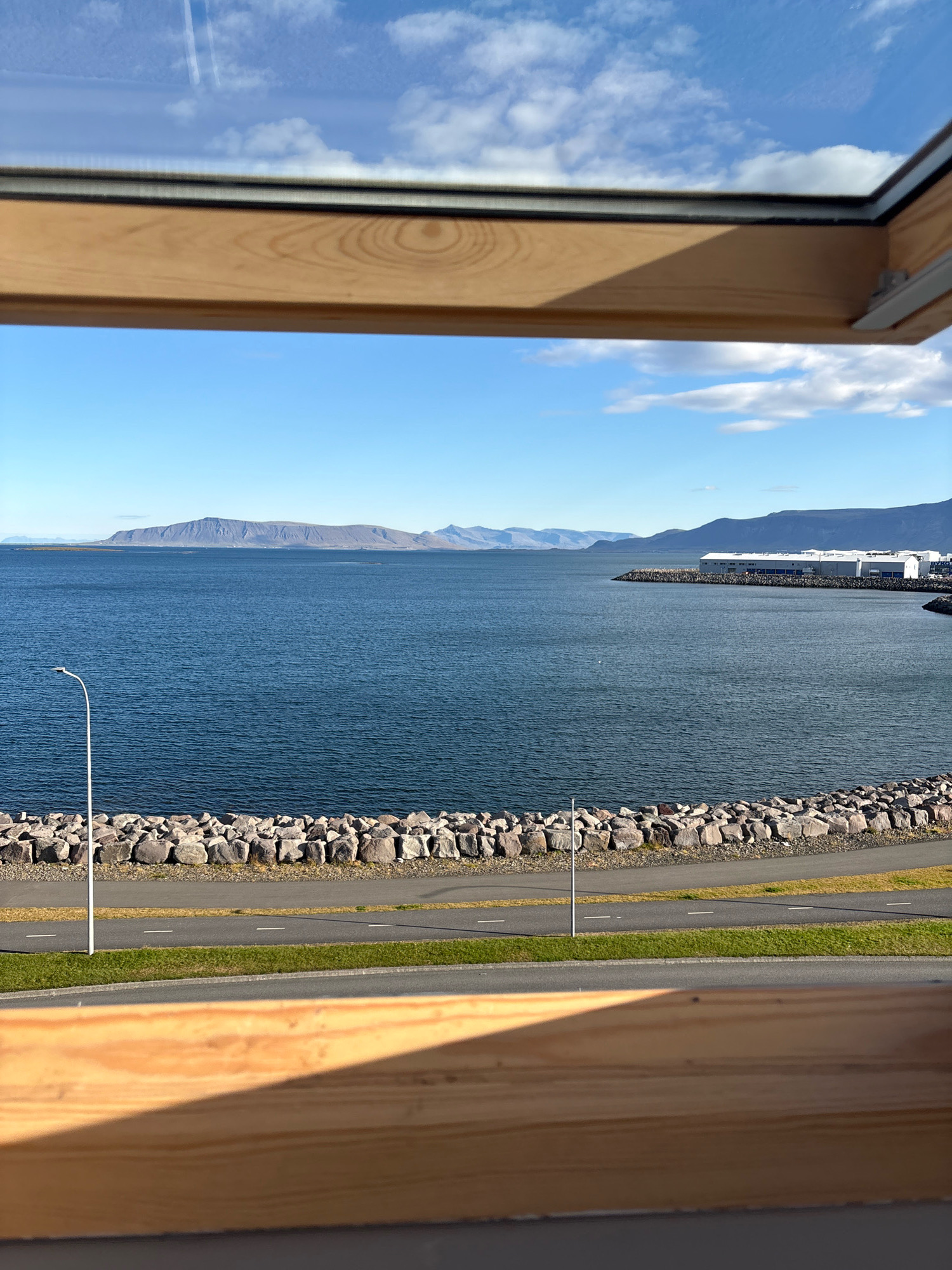 The image shows a view of the ocean and coastline from a room with a wooden window frame. In the foreground, a wooden beam forms the bottom of the frame, casting a shadow. Beyond the window, a body of deep blue water stretches to the horizon. The coastline is lined with a rocky barrier and a paved path, possibly a road or promenade, with a streetlamp visible on the left. In the distance, a long, low-lying mountain range forms the backdrop against a partly cloudy blue sky. On the right side of the image, a harbor with industrial-looking buildings can be seen along the coast. The overall impression is a scenic view of the Reykjavik coastline under a bright, clear sky.