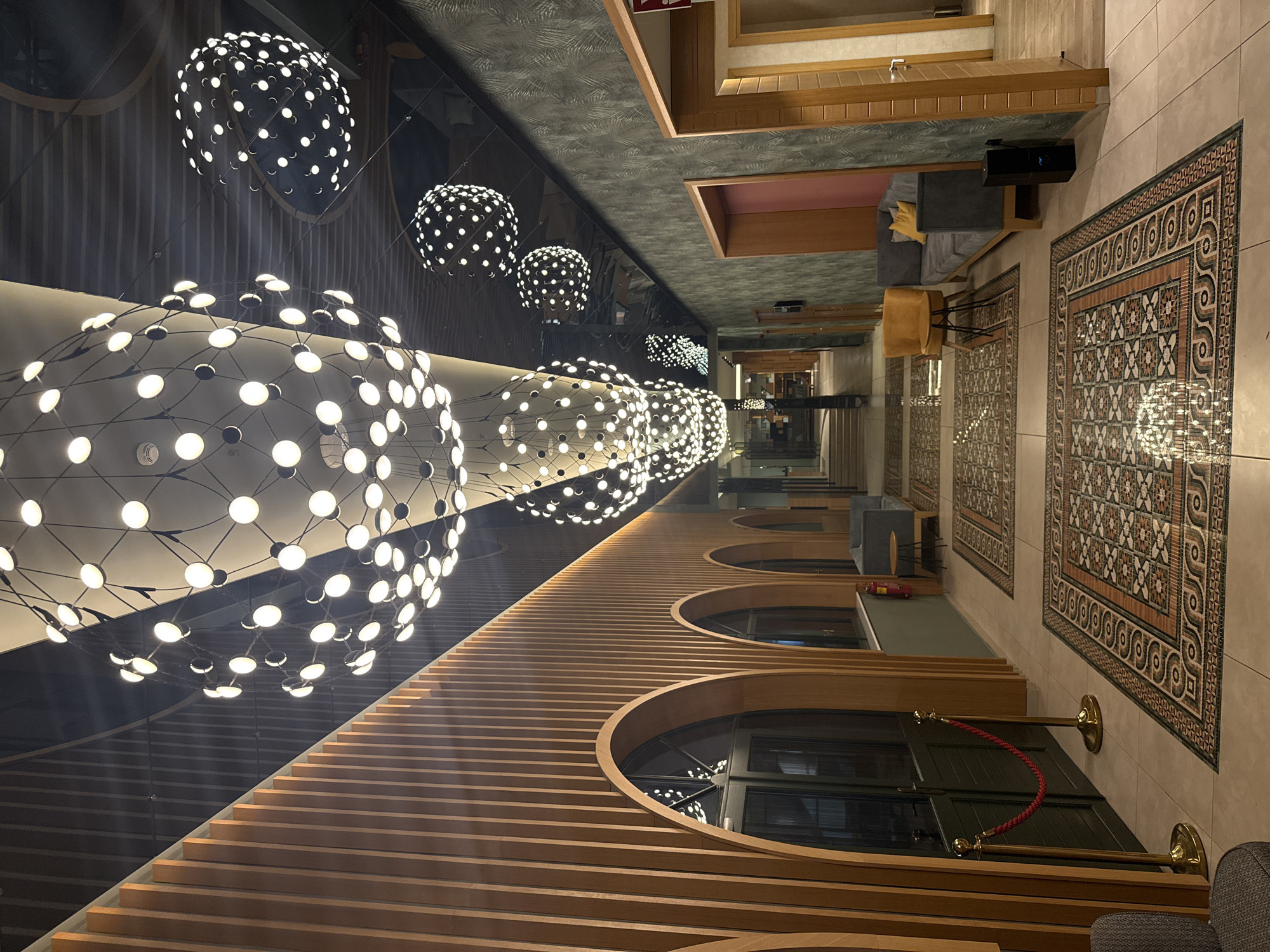This image depicts the interior of a modern building, possibly a hotel lobby or a commercial space, located in the city of Artemida Municipal Unit, Greece. The scene is characterized by a long, well-lit hallway with a high ceiling featuring multiple large, spherical light fixtures composed of many small, bright lights. The walls are a mix of textures and materials, including a section with vertical wooden slats and arched openings, and another section with a dark, textured wallpaper.
The floor is tiled and adorned with several large, ornate rugs with geometric patterns in earth tones. Along the right side of the hallway, there are doorways leading to different rooms. Seating areas are visible, including a grey sofa with yellow cushions and a bright yellow chair placed near a small circular table. On the left side, there are double doors with dark frames and glass panels, and a velvet rope barrier with gold stanchions. The overall ambiance is one of contemporary design and sophistication. No people are visible in the image.