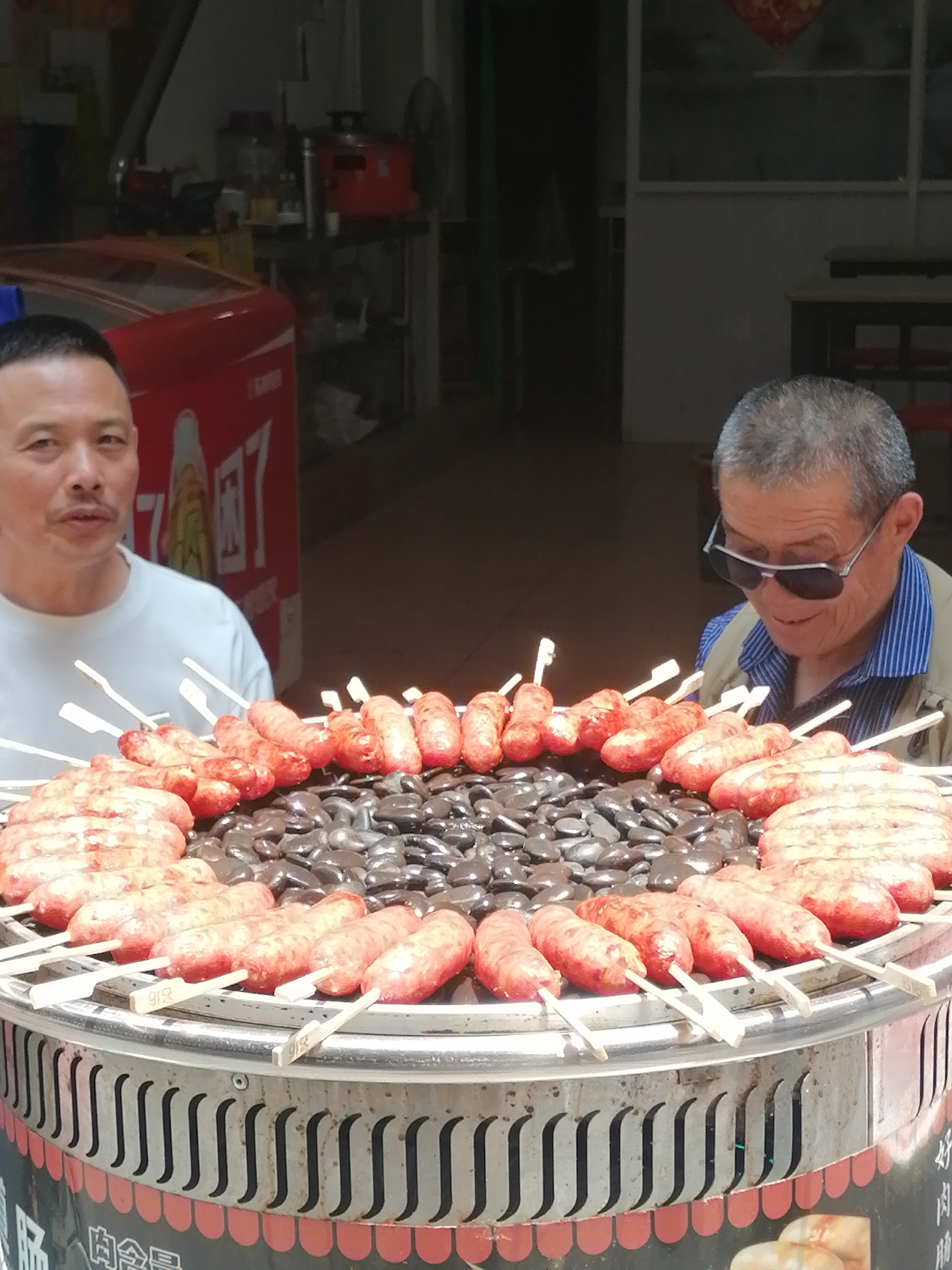 Two men are positioned behind a circular grill adorned with skewers of sausages, situated in Hekou, China. The man on the left wears a white t-shirt, while the man on the right sports sunglasses and a blue striped shirt beneath a light tan jacket. The grill features black stones atop its surface, and the skewers hold evenly spaced sausages. The grill's base is decorated with a scalloped red pattern. Behind the men is a red beverage cooler and the entrance to a building.