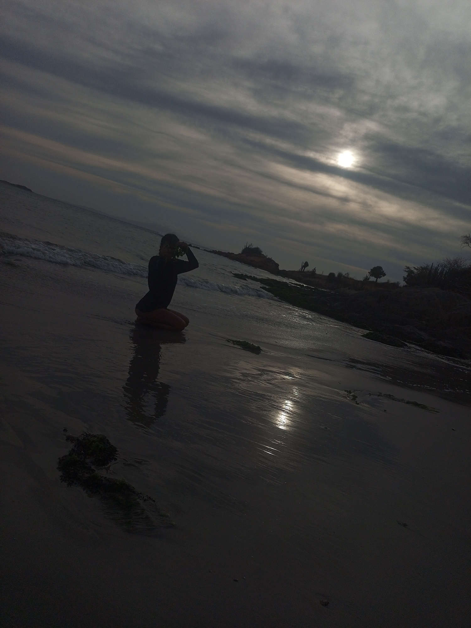 A long shot captures a person kneeling on a sandy beach, their silhouette prominent against the pale horizon. The sun, veiled by a layer of clouds, casts a faint glow across the scene, reflected in the wet sand. Gentle waves approach the shoreline, while vegetation dots the landscape in the distance. The location is Juan Griego, Venezuela.