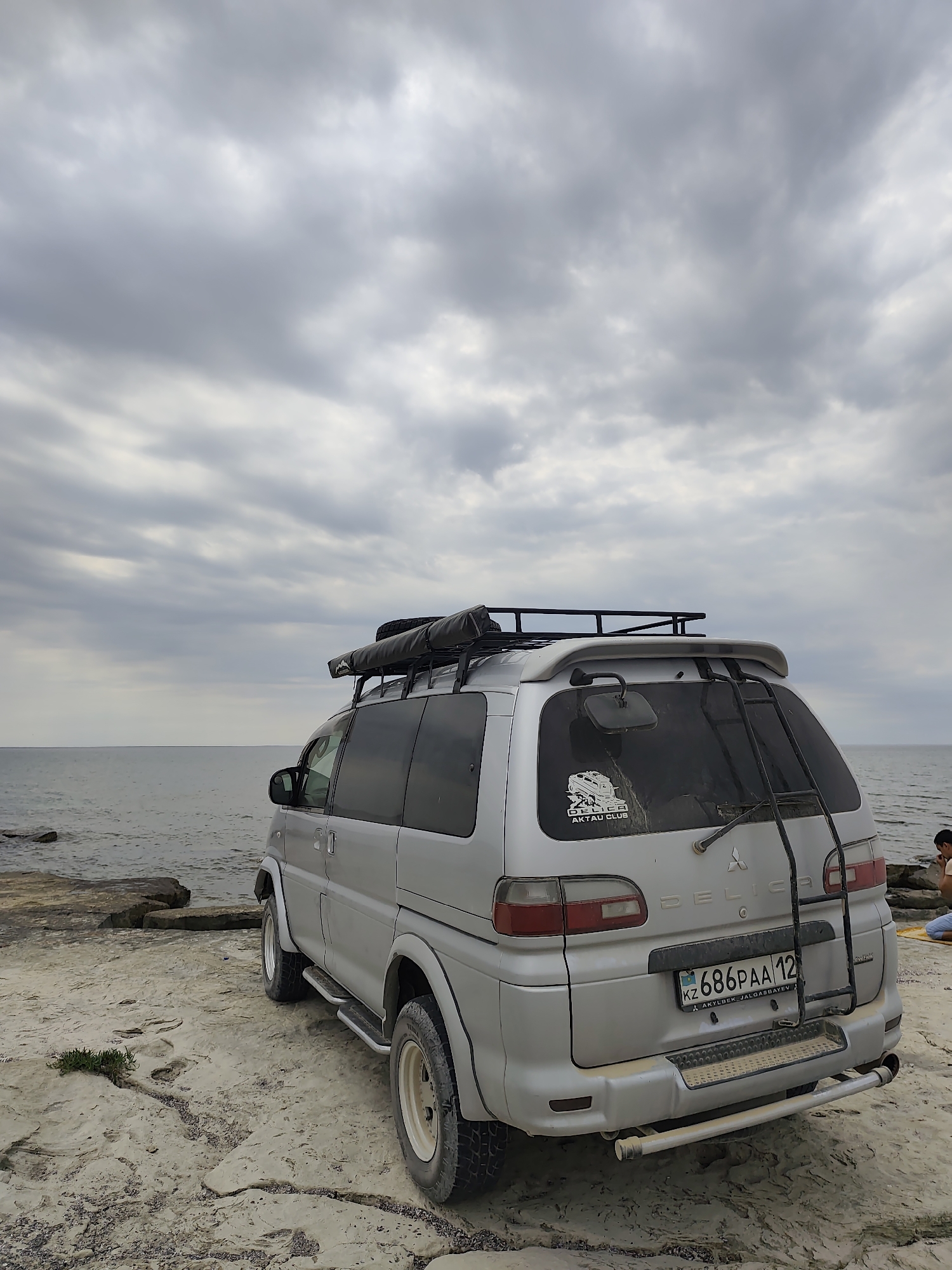 A silver Mitsubishi Delica van is parked on a rocky, flat coastline overlooking the sea. The van has a roof rack with an awning and is facing away from the camera, with its rear visible. A ladder is mounted on the back of the van. A license plate reading "KZ 686PAA 12" is attached to the rear bumper. A sticker on the rear window says "DELICA AKTAU CLUB". The sky is overcast with gray clouds. In the background, to the right, a person is seated on the rocks. The location is identified as Aqshuqyr, Kazakhstan.