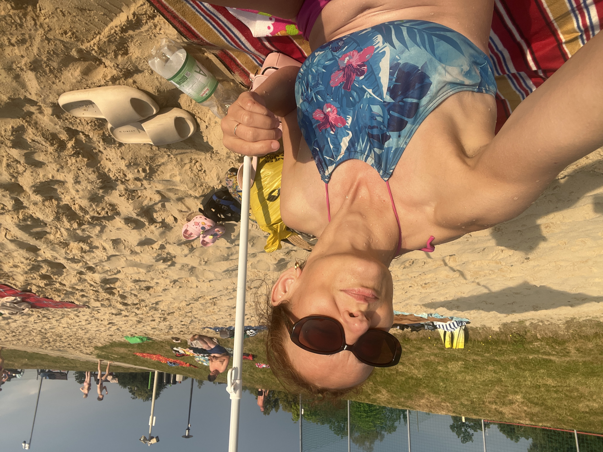 A woman in a blue and pink floral bikini is sitting on a sandy beach, holding a white umbrella pole. She is wearing large, dark sunglasses and has a gold ring on her right hand. To her right, a yellow bag and a pink bag are visible. Several pairs of sandals, including pink and beige slides, are scattered on the sand. A water bottle with a green label lies near the beige slides. In the background, other beachgoers are visible, some sitting on towels and others standing or walking on a grassy slope. The beach is located at a park with a fence and trees in the distance. The lighting suggests it is late afternoon or early evening.