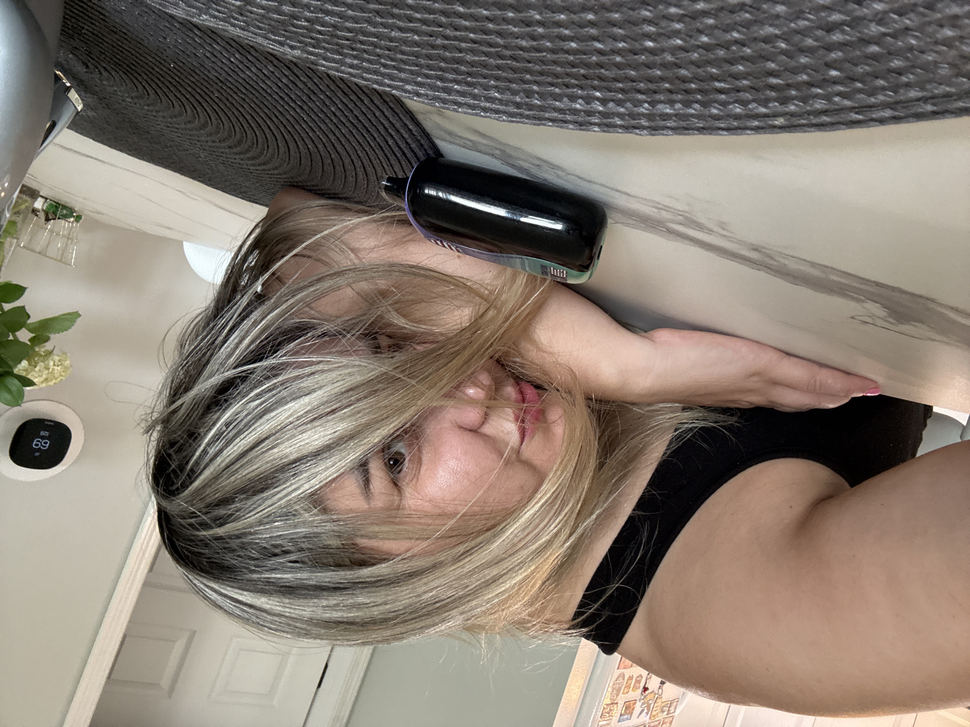 In an indoor setting, a woman with blonde and brown hair lies face down on a marble-patterned table. She is wearing a black tank top. Her head is resting on her arms, and her hair partially obscures her face. A black vaping device is positioned on the table in front of her. A grey placemat is visible underneath her arms and the vaping device. In the background, a white door, a thermostat displaying "69" degrees, and a vase with white flowers are visible. The overall impression is a casual, relaxed moment. The specific location within the city of Wheeling, United States of America, cannot be determined from the image.