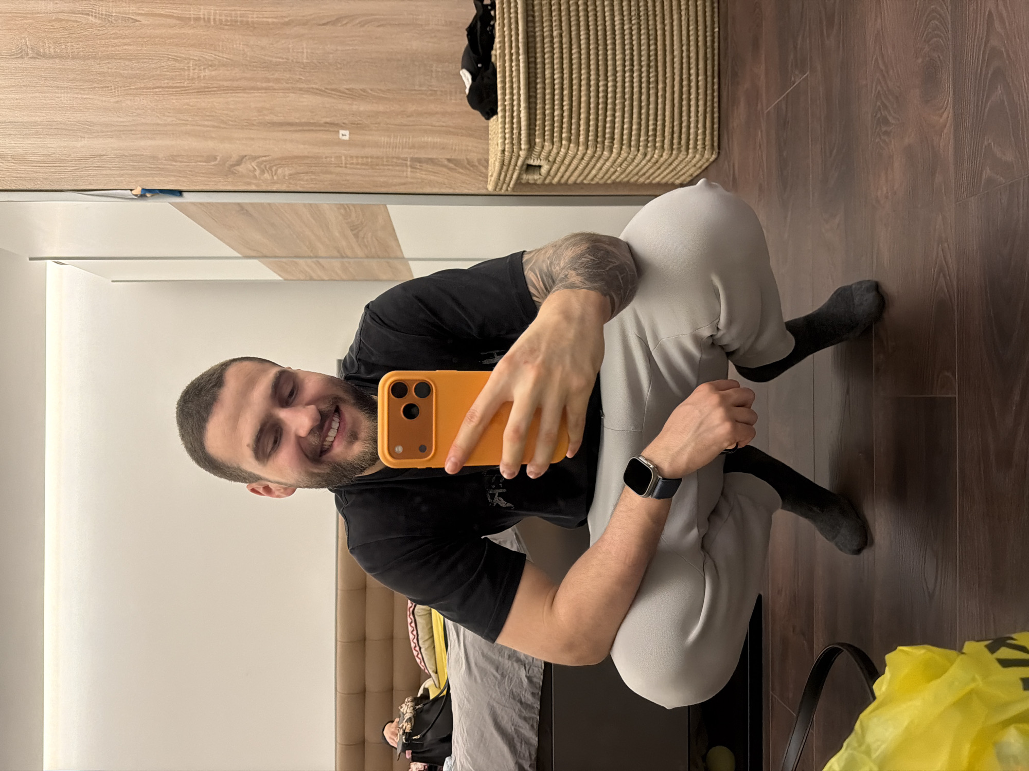 A man is captured in a mirror selfie, with the image rotated 90 degrees counter-clockwise. He is squatting or sitting on the floor, holding an orange-cased smartphone with a visible triple-lens camera module in his right hand. He is smiling broadly, revealing his teeth, and has short dark hair and a beard. He wears a black t-shirt, light grey sweatpants, and dark grey socks. An intricate tattoo is visible on his right forearm/bicep, and he wears a smartwatch with a dark strap on his left wrist.
The background, reflected in the mirror, shows an indoor setting, likely a modern bedroom or living space in Sofia, Bulgaria. Elements visible include a light wood-grained wardrobe or cabinet, a woven basket next to it, and a dark wooden floor. Part of a bed or headboard with light-colored upholstery is also visible. The lighting is bright, suggesting daytime. In the lower right corner, a yellow plastic bag is visible, with the word "KAUFLAND" printed in black letters. A dark bag or purse strap is also partially seen near the yellow bag. A small, unreadable white rectangular sticker is on the wooden cabinet. The man appears happy and engaged in taking the photograph.