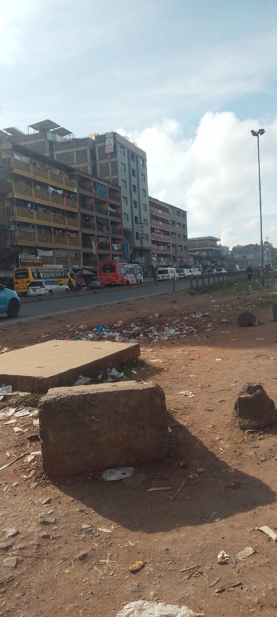 The image depicts a street scene in Nairobi, Kenya, characterized by a mix of residential buildings, commercial vehicles, trash, and undeveloped land. The buildings, appearing to be apartment complexes, vary in height and age, with some showing signs of wear and tear. Clothes are hanging from some of the balconies. A variety of buses and vans are lined up along the side of the street. In the foreground, there is a dirt area cluttered with trash, large concrete blocks, and debris. A street lamp is visible along the road. The sky is partly cloudy.