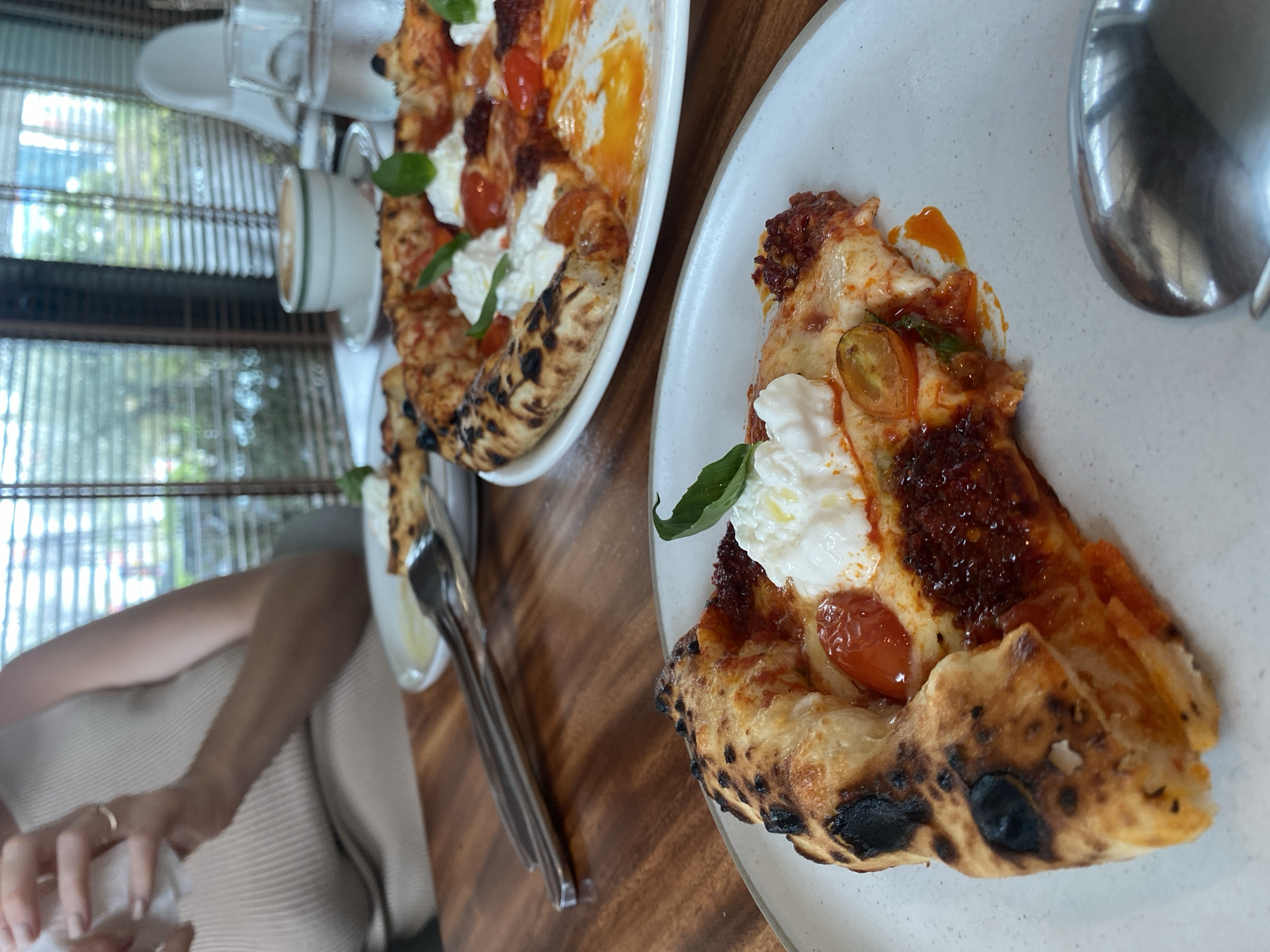 The image captures a close-up of a partially eaten pizza on a white plate, set on a wooden table. A second, similarly topped pizza is visible in the background, along with cutlery, glassware, and a cup. The pizza features a charred crust, tomato sauce, cherry tomatoes, and a dollop of creamy white cheese, garnished with a basil leaf. A person's arm is visible in the lower left corner of the frame. The scene is illuminated by natural light, filtered through Venetian blinds, which suggests an indoor setting in Choeng Thale, Thailand.