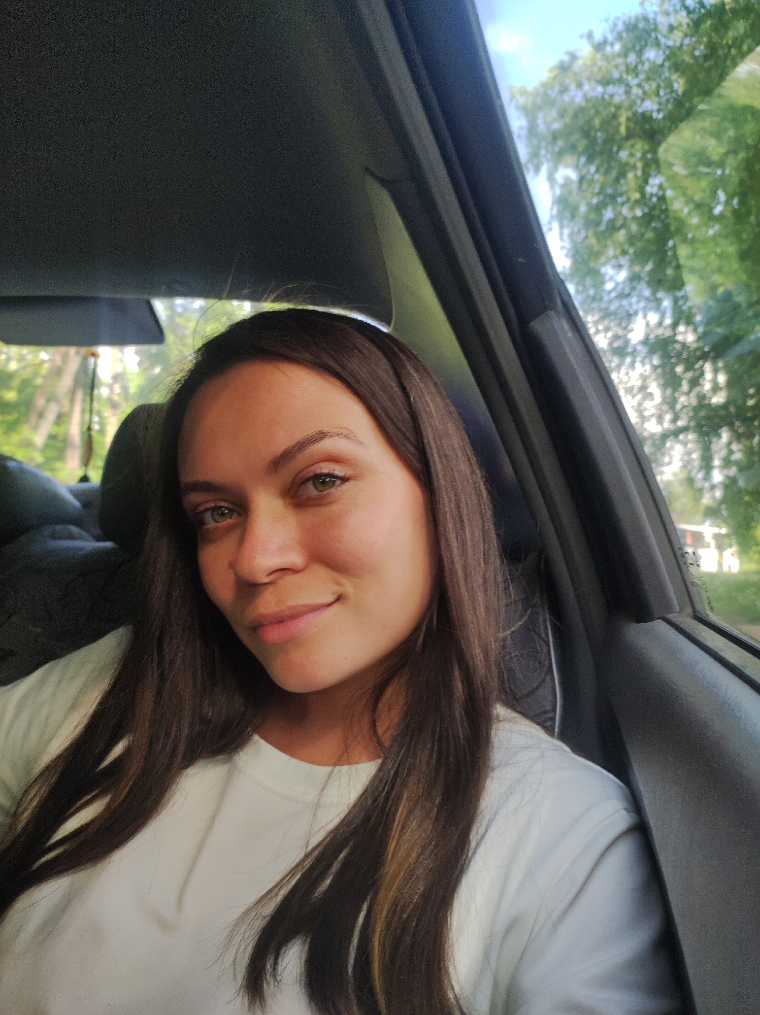 A woman with long, dark brown hair and fair skin is sitting in a car. She is wearing a white t-shirt. She is looking directly at the camera with a slight smile. The car window is open, and trees and a building can be seen outside. The location is Chaikovsky, Russia. The interior of the car is gray and black. A black headrest is visible behind her. A wooden object, possibly a small decorative item, is hanging from the headrest. The sky is partially visible through the trees. The lighting suggests it is daytime. The scene is a selfie taken inside a parked car.