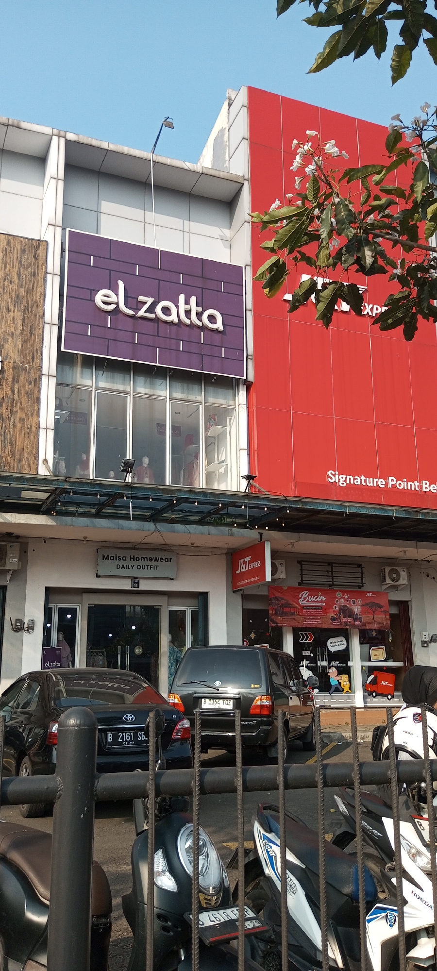 A daytime, eye-level, medium shot captures the exterior of a commercial building complex in Lambangjaya, Indonesia. The building's facade features a prominent red section on the right, displaying the text "Signature Point Bel..." in white cursive font. To the left, a large purple sign with white lettering reads "elzatta." Below these signs, a covered walkway with hanging lights spans the width of the building, leading to various storefronts.
Several vehicles are parked in front of the shops, including a black sedan with license plate B 2161 SB and a black SUV with license plate B 1164 TGZ. Motorcycles are also visible, some partially obscured by a metal barrier in the foreground. Storefronts include "Maisa Homewear DAILY OUTFIT" with a number "11" below it. A red and white J&T Express sign is also present. The sky is a clear, bright blue, and green foliage from a tree with white flowers hangs into the frame from the upper right. The overall impression is that of a bustling commercial area on a sunny day.