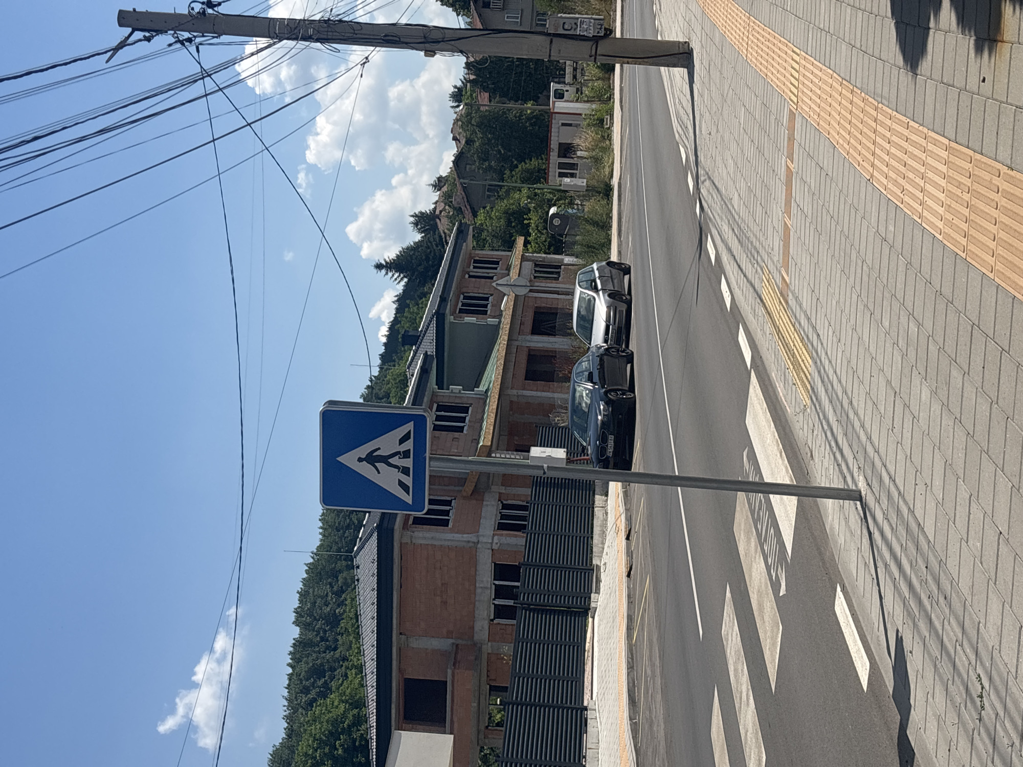 This image shows a street scene with a pedestrian crossing and construction. The scene is outdoors during the day under a clear blue sky with some fluffy white clouds. The sun casts shadows across the scene, indicating it is likely daytime.
The main subjects are a pedestrian crossing sign and two cars on the street. The pedestrian crossing sign is a blue square with a white triangle inside, depicting a person walking. The cars are a silver SUV and a black sedan parked alongside the road.
In the background, there is a multi-story building under construction, with exposed brickwork and framing. The building appears to be in the early stages of construction, with open window frames and no finished facade. Behind the construction, there are trees and a hillside, suggesting a semi-urban or suburban setting. A utility pole with numerous overhead wires is also prominently featured on the left side of the image.
The sidewalk adjacent to the street has a tactile paving strip, indicated by the textured orange tiles, which are often used to guide visually impaired pedestrians. The street itself has clear white markings for the pedestrian crossing.
No specific activity or event is clearly taking place, other than the presence of parked cars and the ongoing construction. The overall mood is one of everyday life with ongoing development. There is some text visible on the license plate of the black car, but it is not legible.