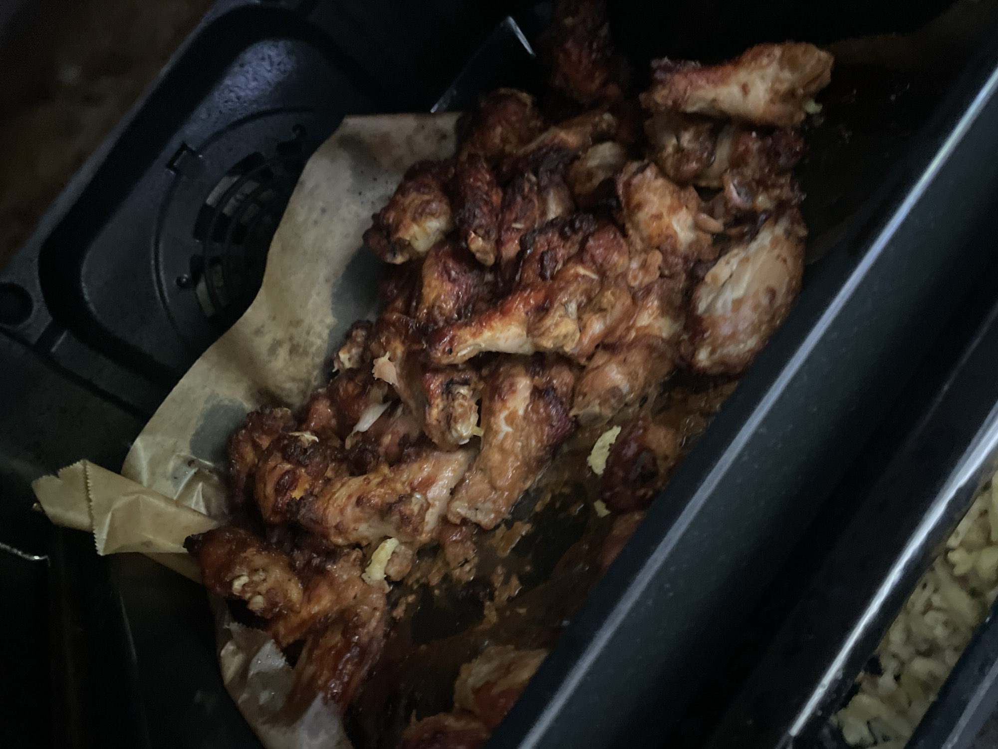 The image shows cooked chicken wings in a black container lined with parchment paper. The wings appear to be heavily seasoned and possibly fried or baked. The container seems to be part of a larger tray, with a glimpse of another compartment filled with a light-colored substance, possibly cooked pasta or potatoes. The location of Wienhausen, Germany cannot be validated from the image data.