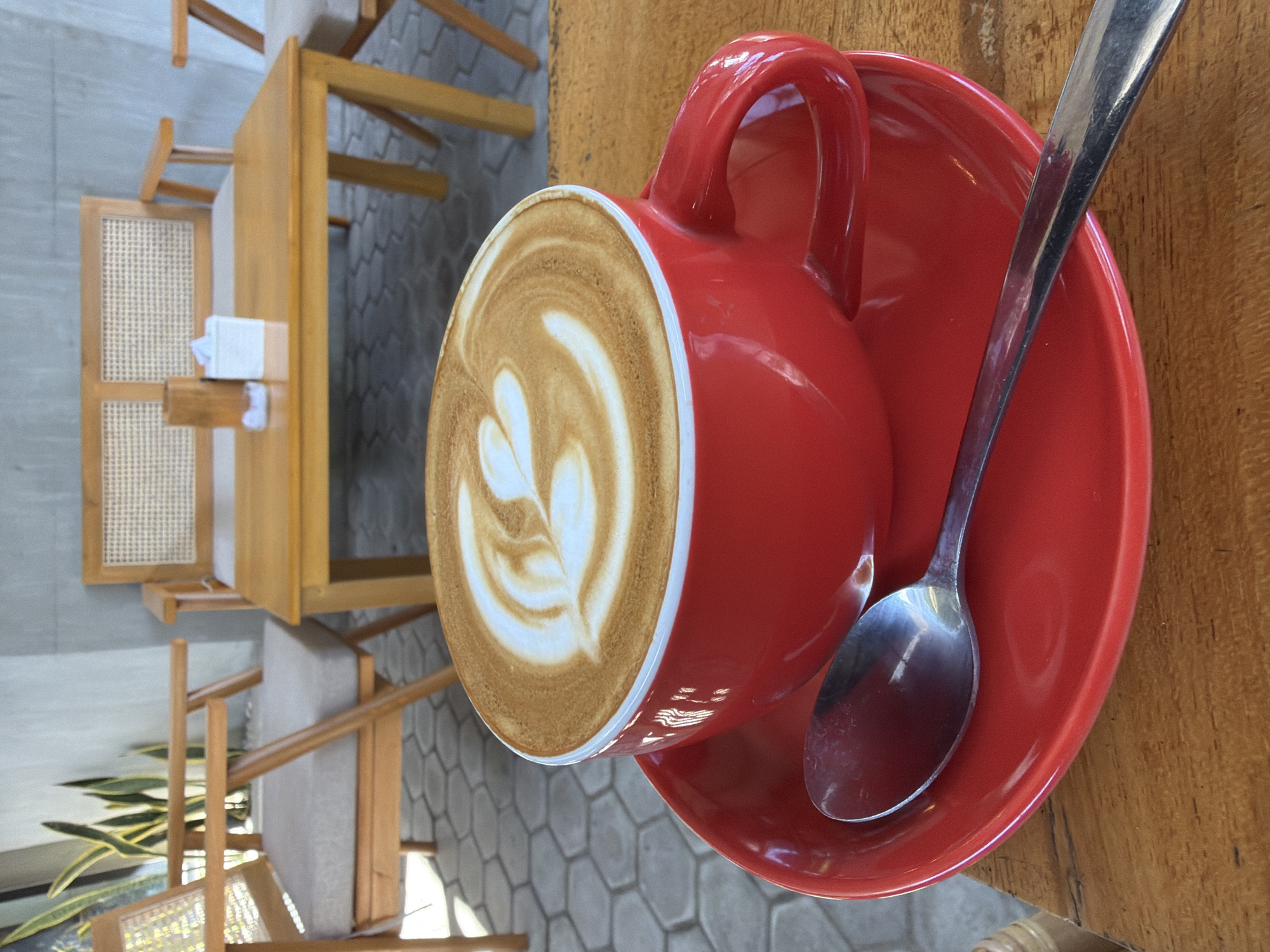 A red ceramic coffee cup with latte art sits on a matching red saucer, which is placed on a wooden table. A silver spoon rests on the saucer. In the background, there are wooden chairs with woven seats and a wooden table with a tissue dispenser. The scene appears to be outdoors, possibly at a cafe in Kuta, Indonesia, with hexagonal-patterned paving visible on the ground.